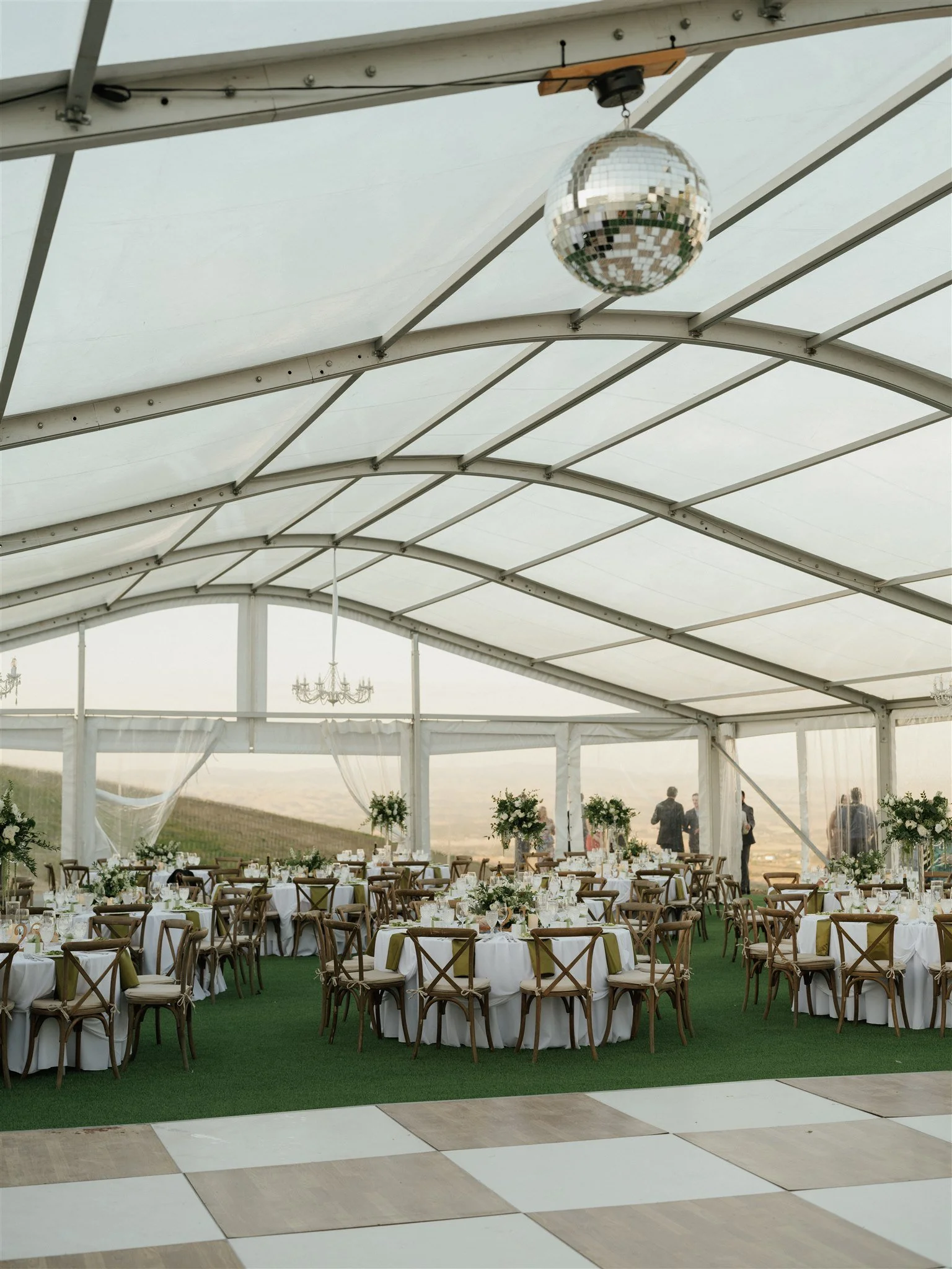 Wedding reception setup inside a large white tent with round tables covered in white tablecloths, decorated with floral centerpieces, surrounded by wooden chairs, with a green carpet and a checkered dance floor in the foreground.