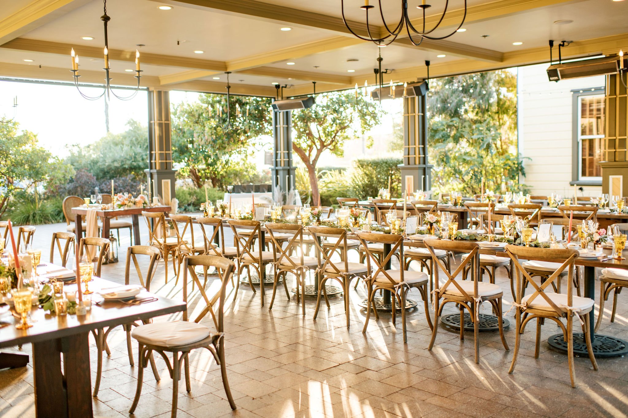 An elegantly decorated outdoor dining area with wooden tables and chairs, set with floral centerpieces, candles, and glassware, under a covered patio with hanging chandeliers and large windows revealing lush greenery outside.