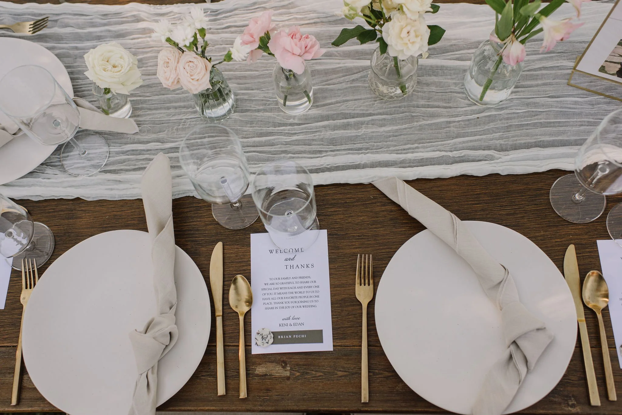 Table setting with white plates, gold utensils, beige cloth napkins, wine glasses, floral centerpieces with roses and lisianthus in glass vases, wedding program, and a striped table runner.