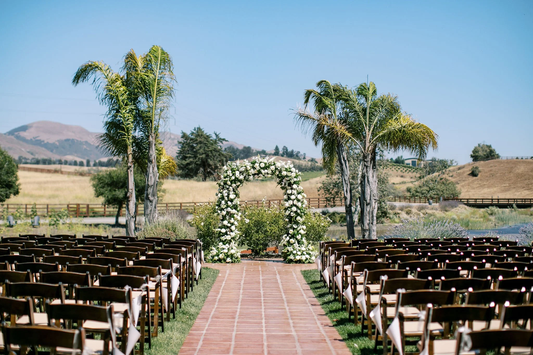 Outdoor wedding ceremony setup on a brick pathway with chairs on both sides, an arch decorated with white flowers, palm trees, green grass, and a scenic rural landscape with rolling hills in the background.