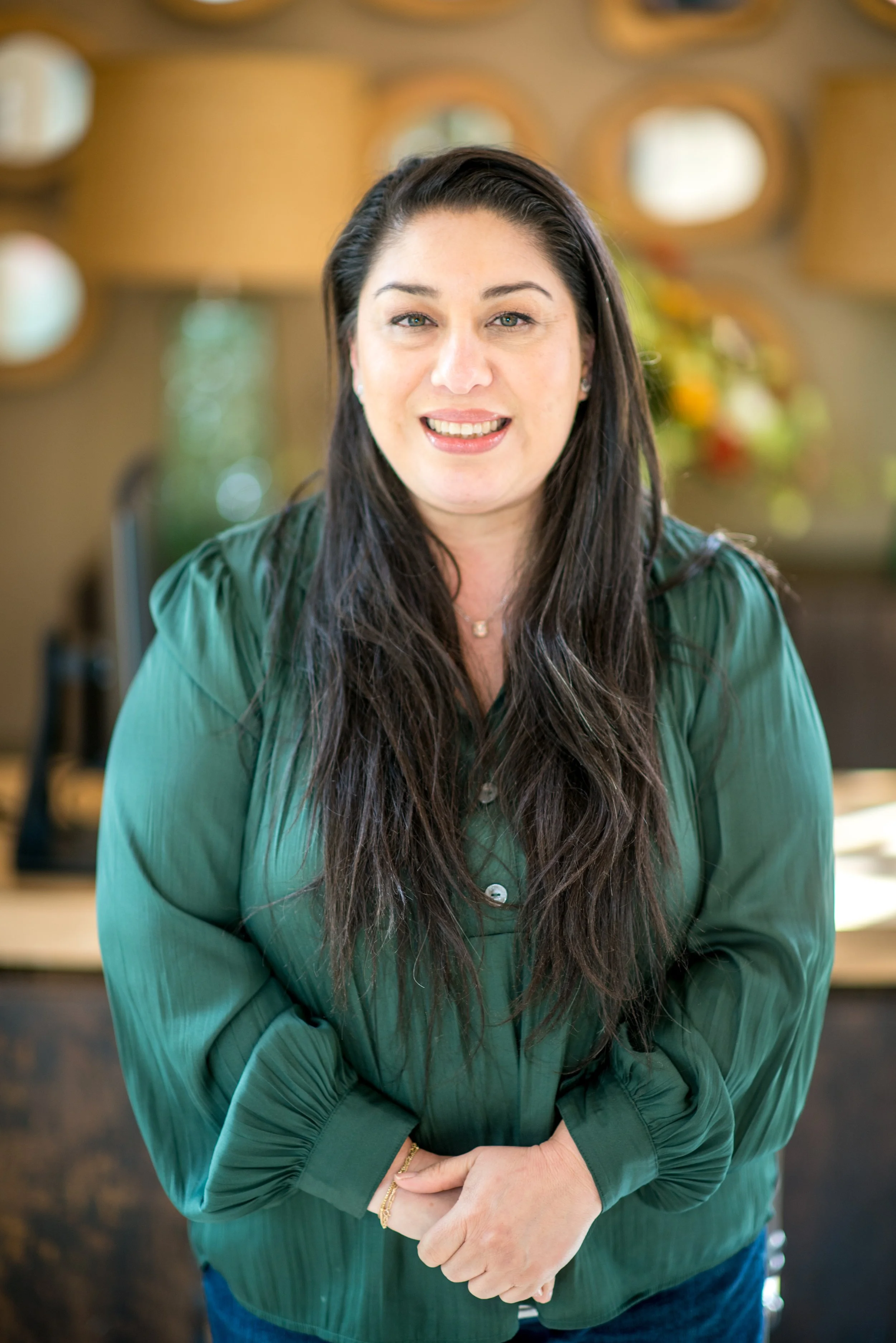 A woman with long dark hair wearing a green silky blouse, standing indoors with a blurred background of decorative wall mirrors and colorful flowers.