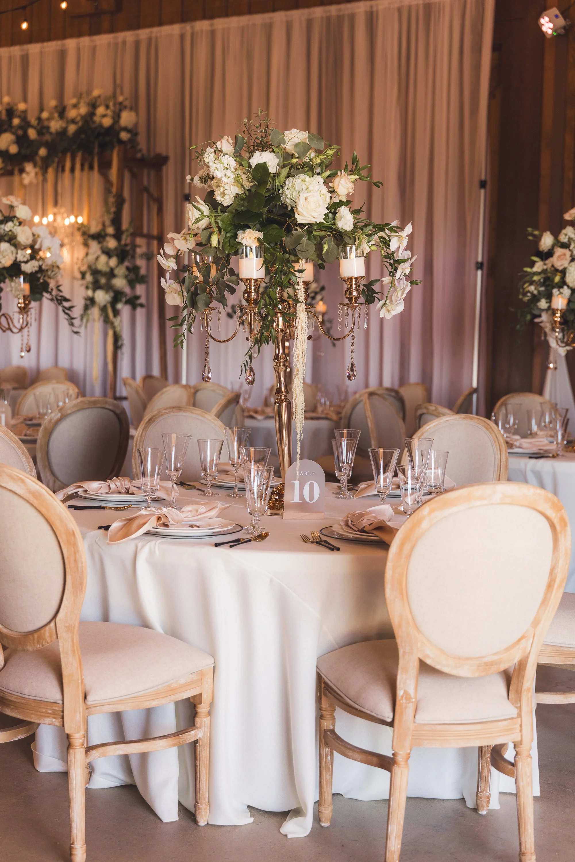 Wedding reception table decorated with a tall floral centerpiece, candelabras, and glassware, with a table number '10' sign.