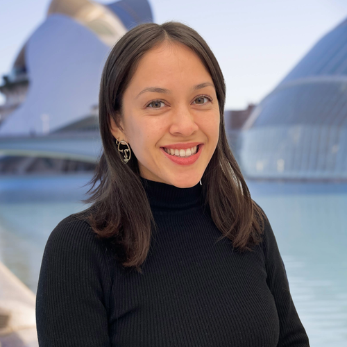 Lola Reynolds, Market Analyst at Outman Consulting, smiling warmly in a black turtleneck with geometric earrings, standing in front of a modern architectural background with reflective water.