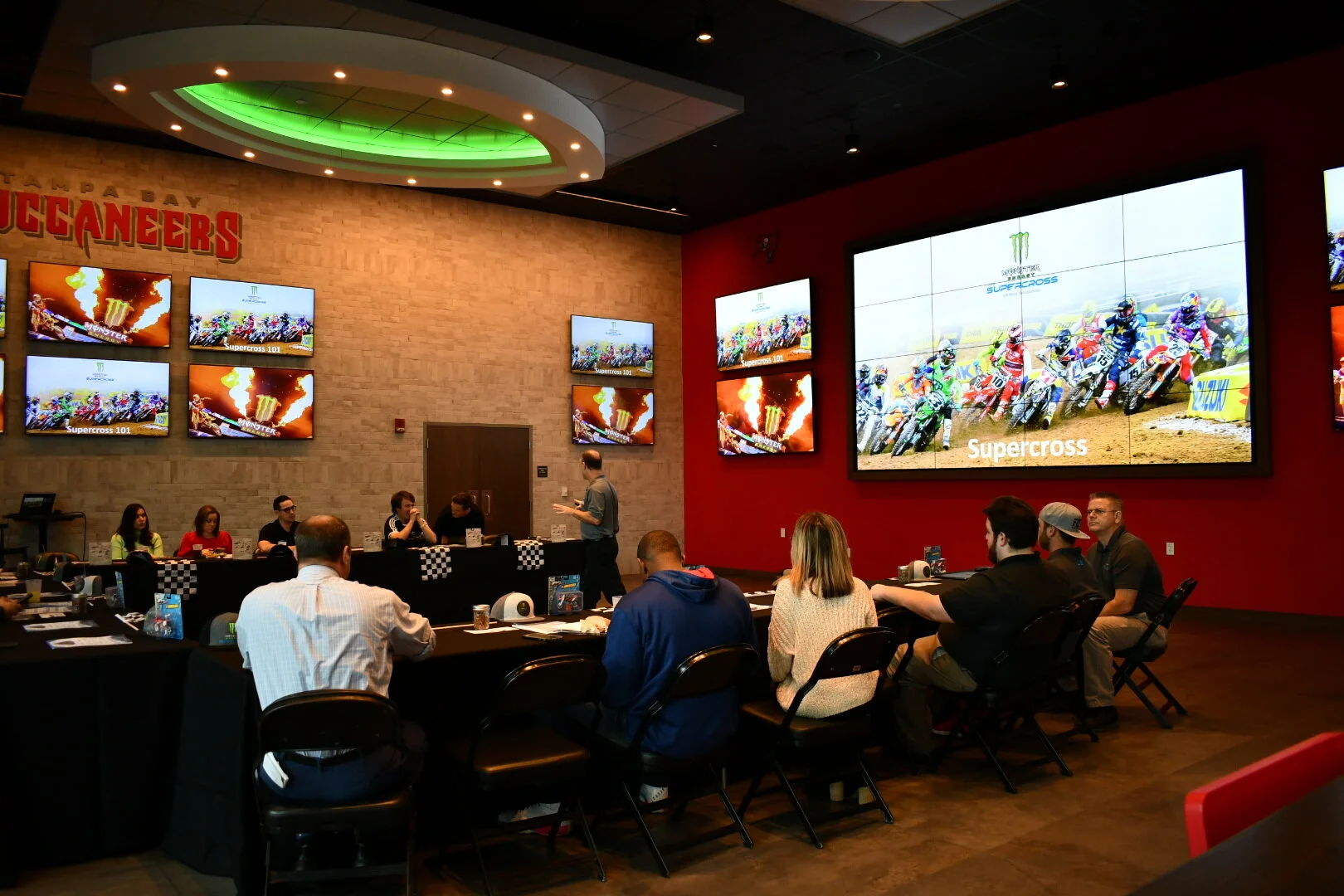 People sit around a U shapped table watching a large screen TV at Bar 76.