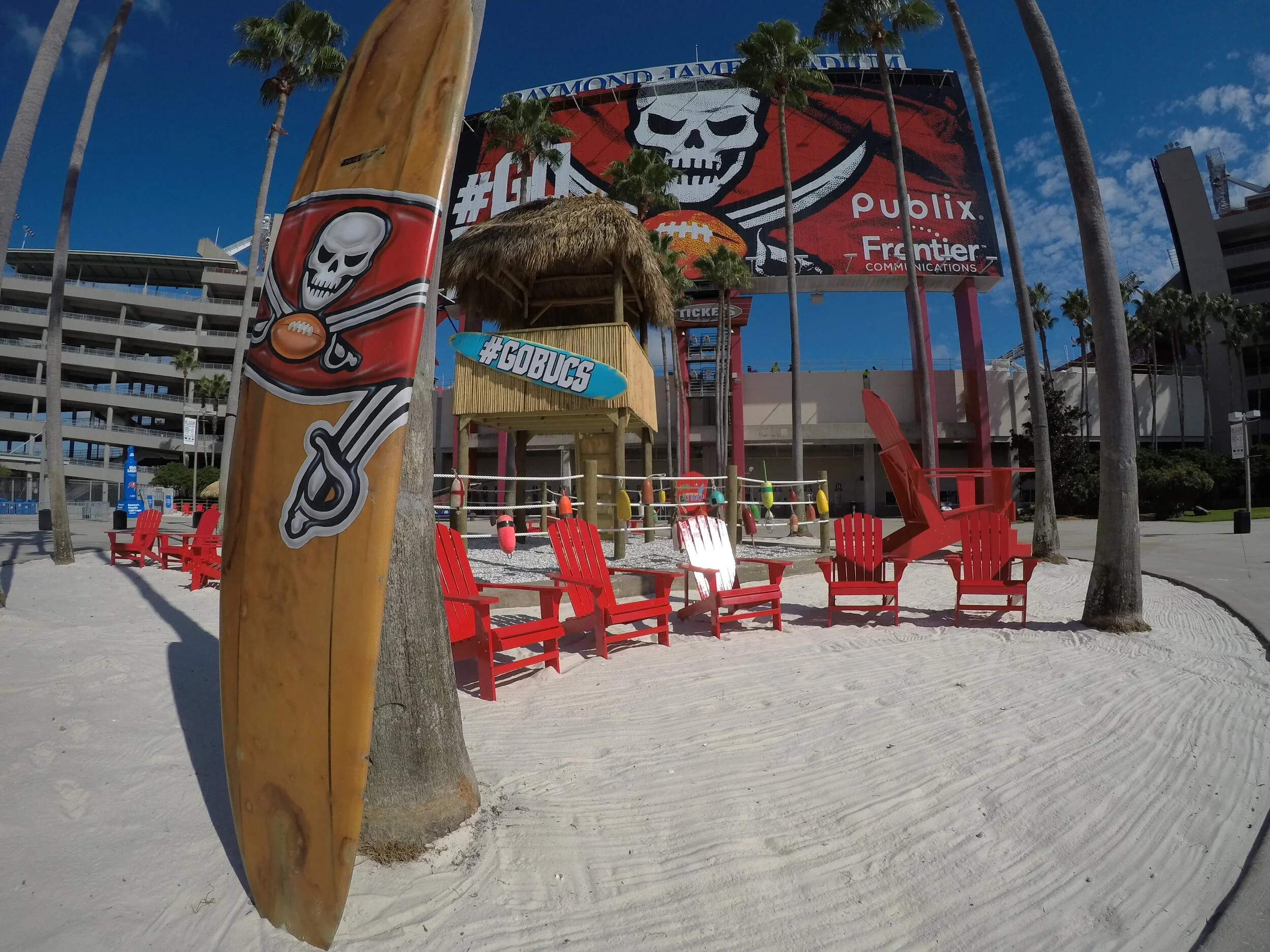 A surfboard with the Buccaneers logo on it is displayed near read beach loung chairs in the sand. 