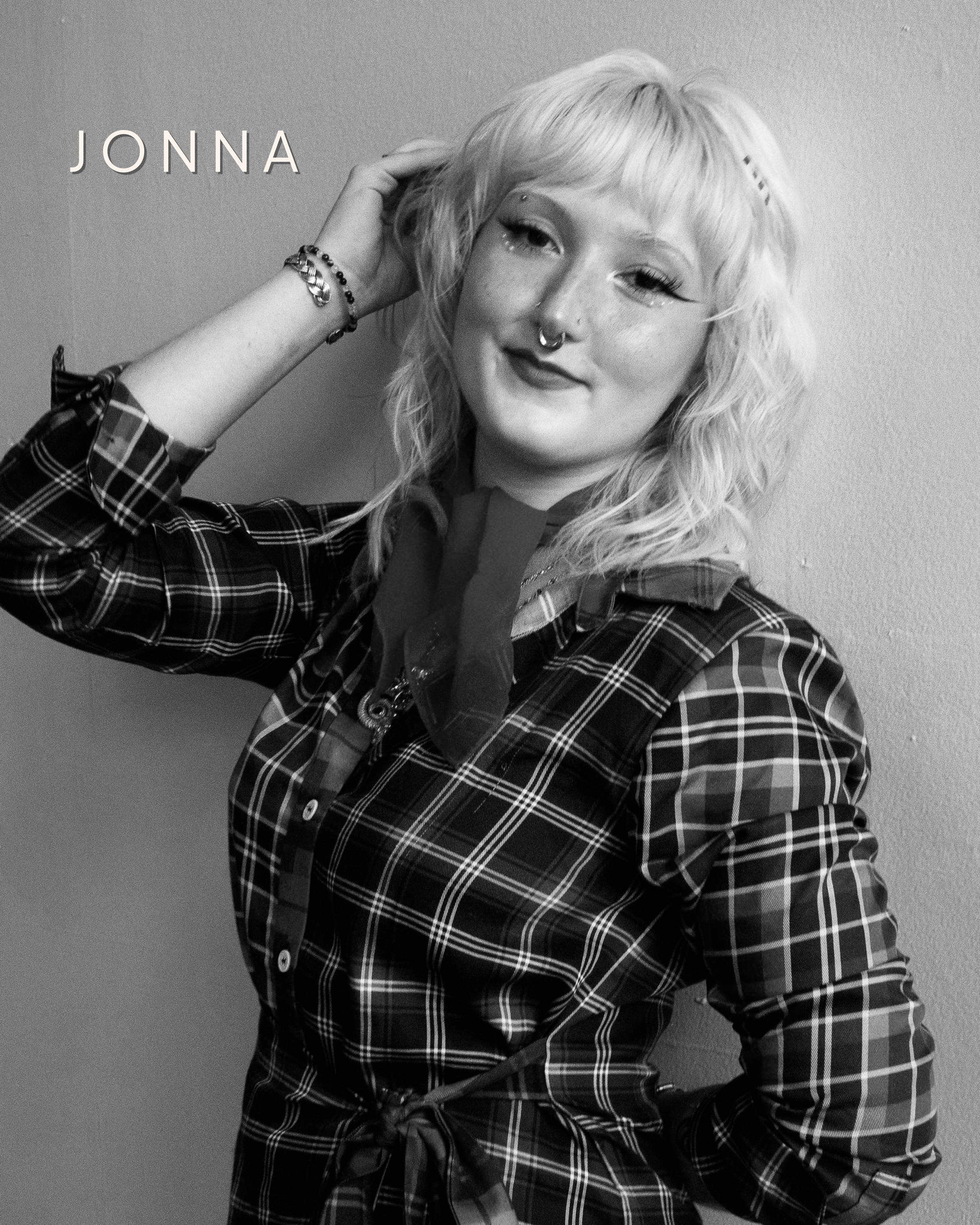 Black and white portrait of a young woman with long dark hair, wearing a light-colored sweater with a dark shirt underneath. She has a septum piercing and is smiling slightly. The background is blurred and dark. The word "JONNA" appears in the upper left corner.