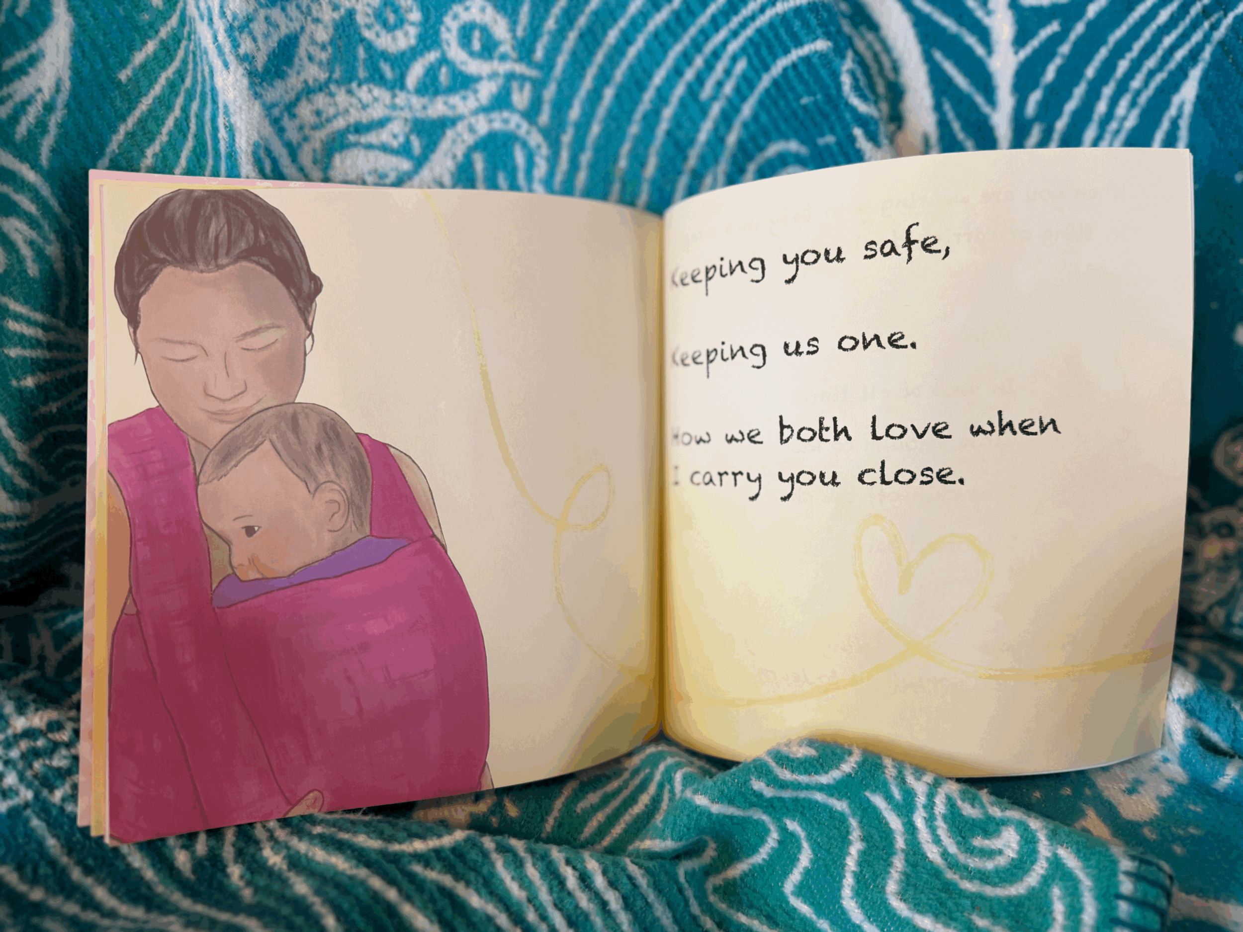 An open illustrated book showing a woman holding a young child in a pink wrap, with the woman’s eyes closed and the child resting on her chest. The right page contains the handwritten text: 'keeping you safe, keeping us one, how we both love when I carry you close.' There is a yellow heart drawing on this page.