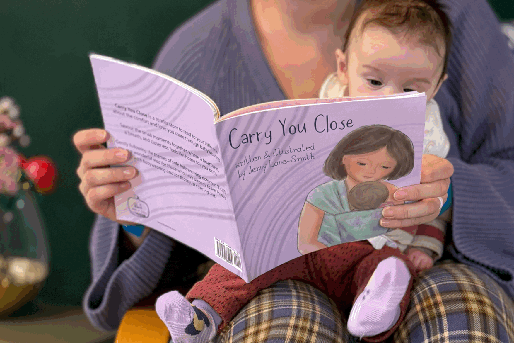 Child sitting on an adult's lap, reading a children's picture book titled 'Carry You Close' with an illustration of a woman wearing her baby in a wrap carrier.