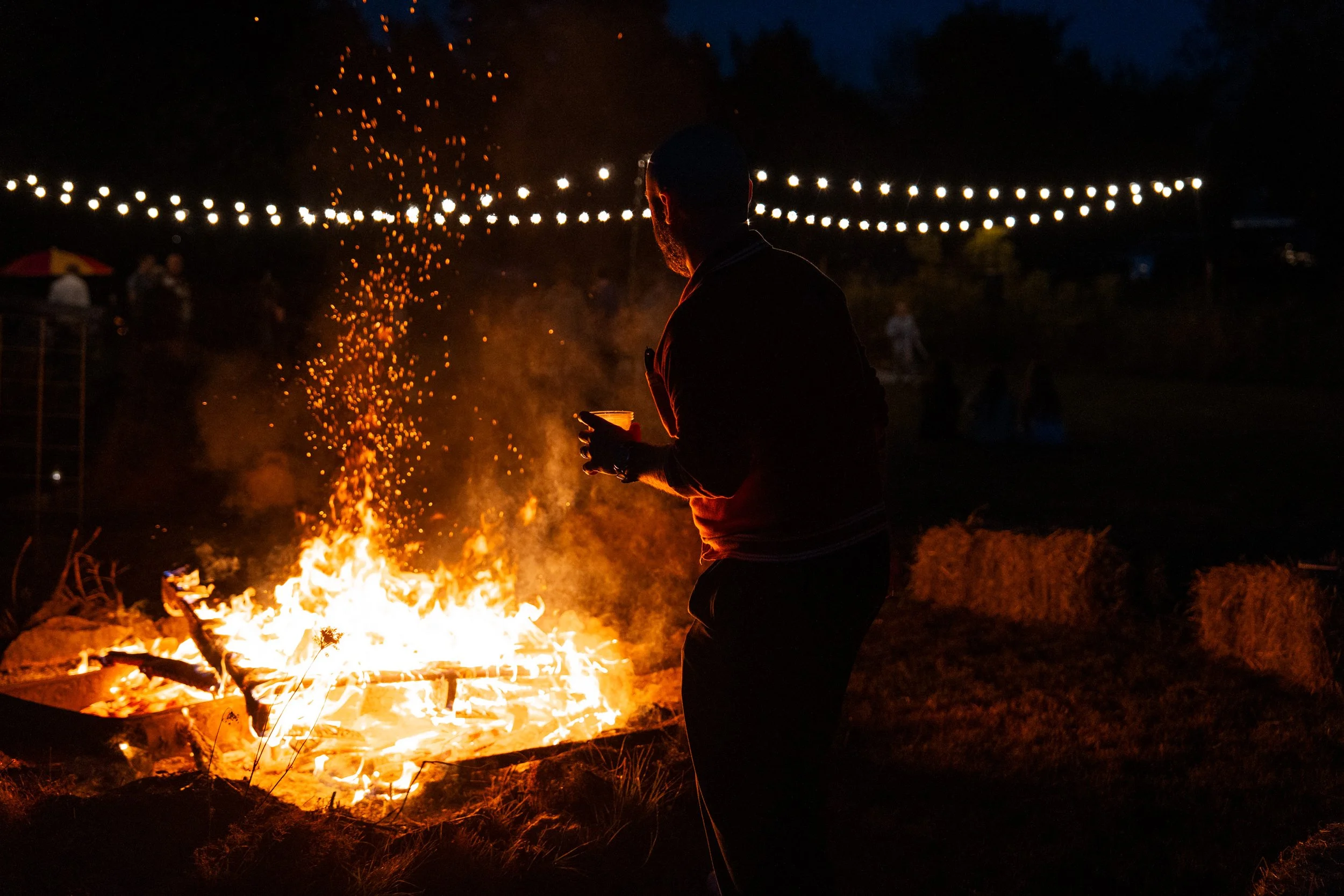 Photo of a bonfire at night with fairy lights in the background.