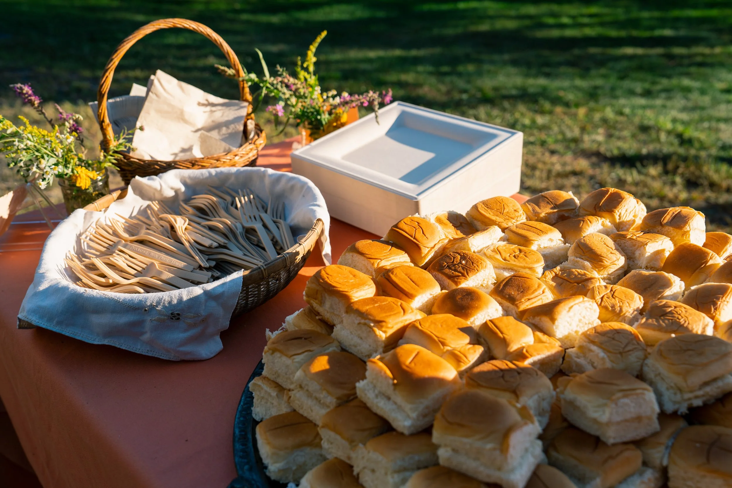 Table with various cutlery, napkins, plates, and Hawaiian rolls outdoors