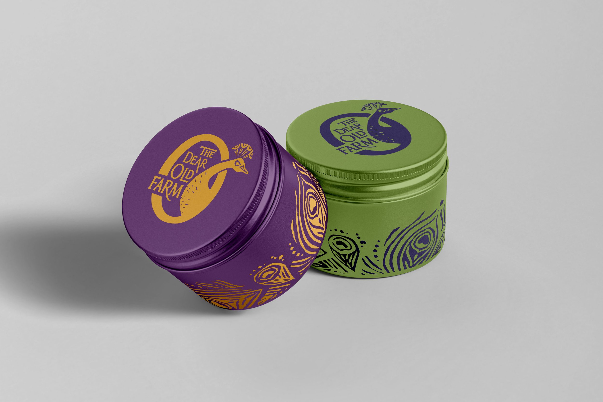 Mockup of two tins, one purple with a yellow Dear Old Farm logo and peacock feather patterns, and one green one with identical markings in a navy blue.