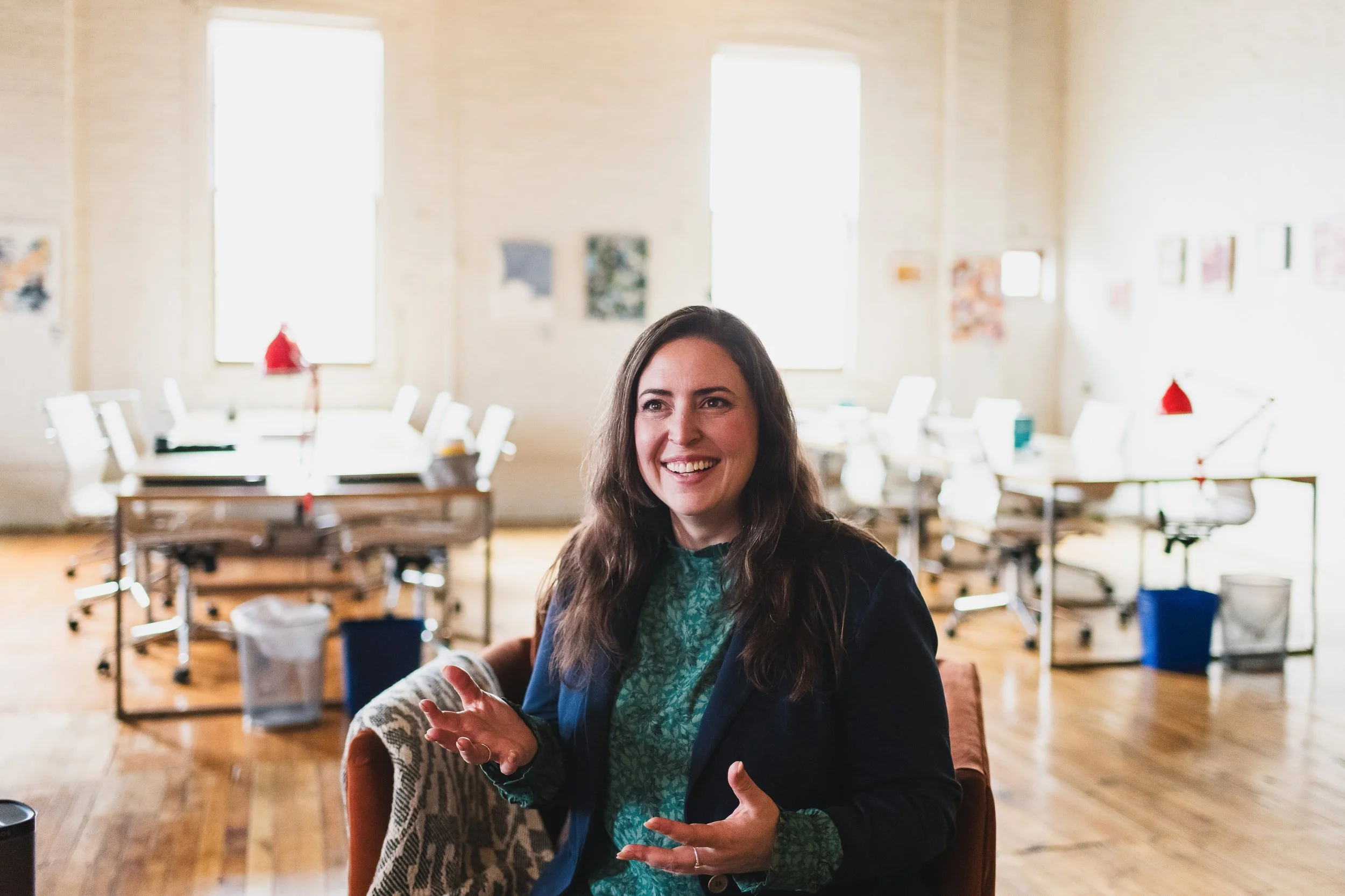 Woman with long brown hair smiling and talking in a bright, modern office with white chairs, desks, and large windows in the background.