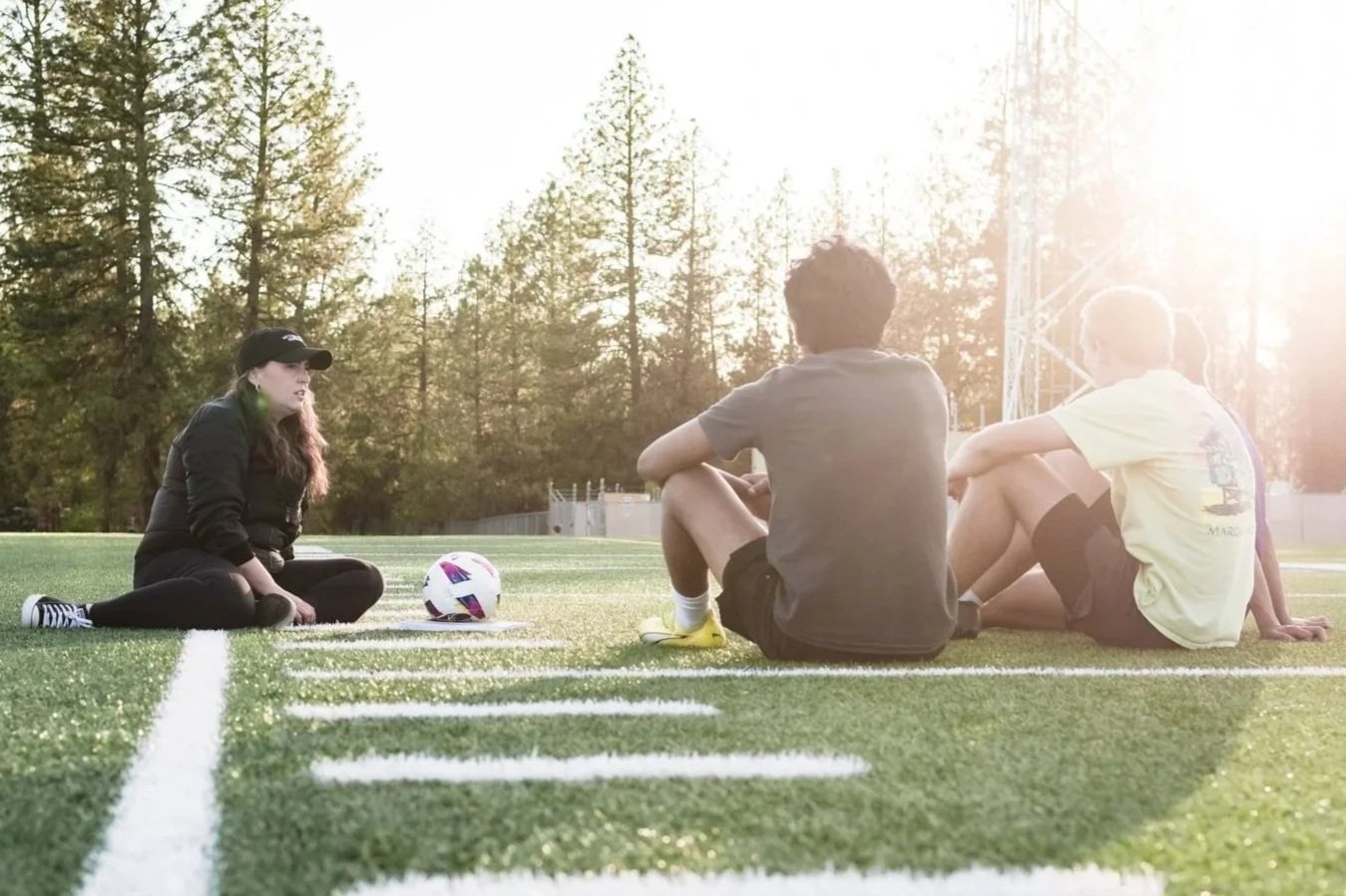 A woman sitting on a soccer field talking to three young men sitting on the ground during late afternoon sunlight.