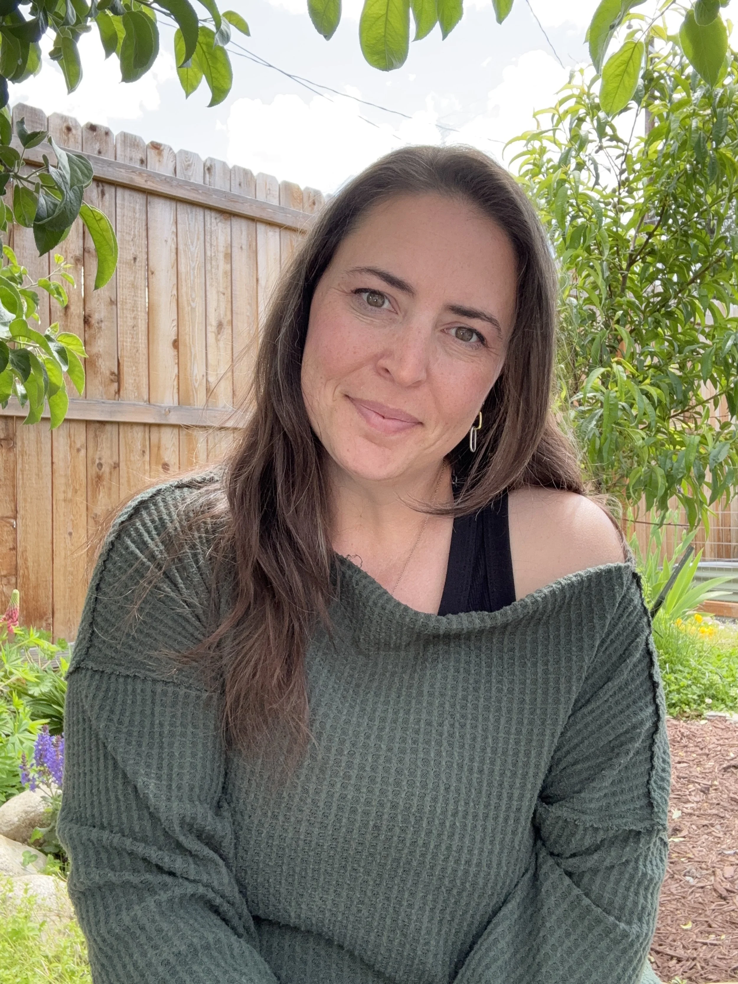 Photo of Stephanie High, founder of Kaizen Catalyst. A woman with long brown hair, wearing a dark tank top and an oversized green sweater, sitting outdoors in a garden with a wooden fence, green leaves, and colorful flowers in the background.