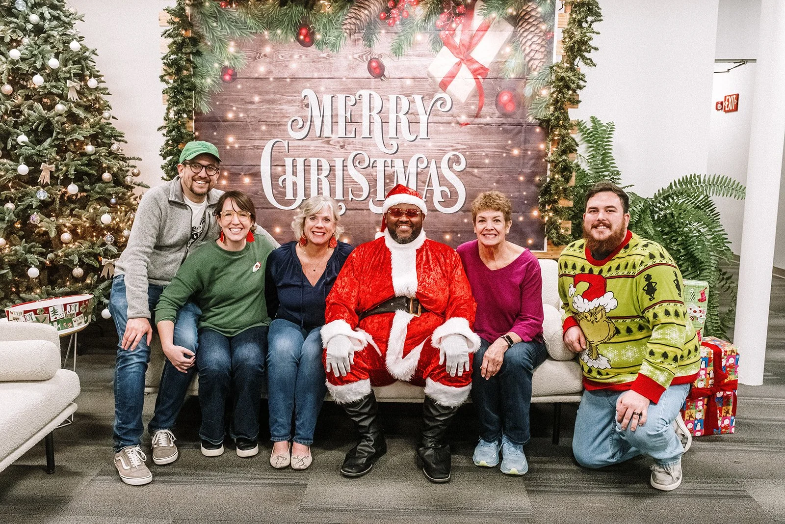Scott, Raegan, Sheryl, Santa, Mo, and Jack pose for a photo in front of a backdrop that says Merry Christmas!