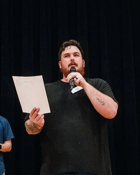 A man with dark hair, a full beard, and light skin, smiling outdoors in front of a brick building and greenery, wearing a maroon t-shirt with a small logo.