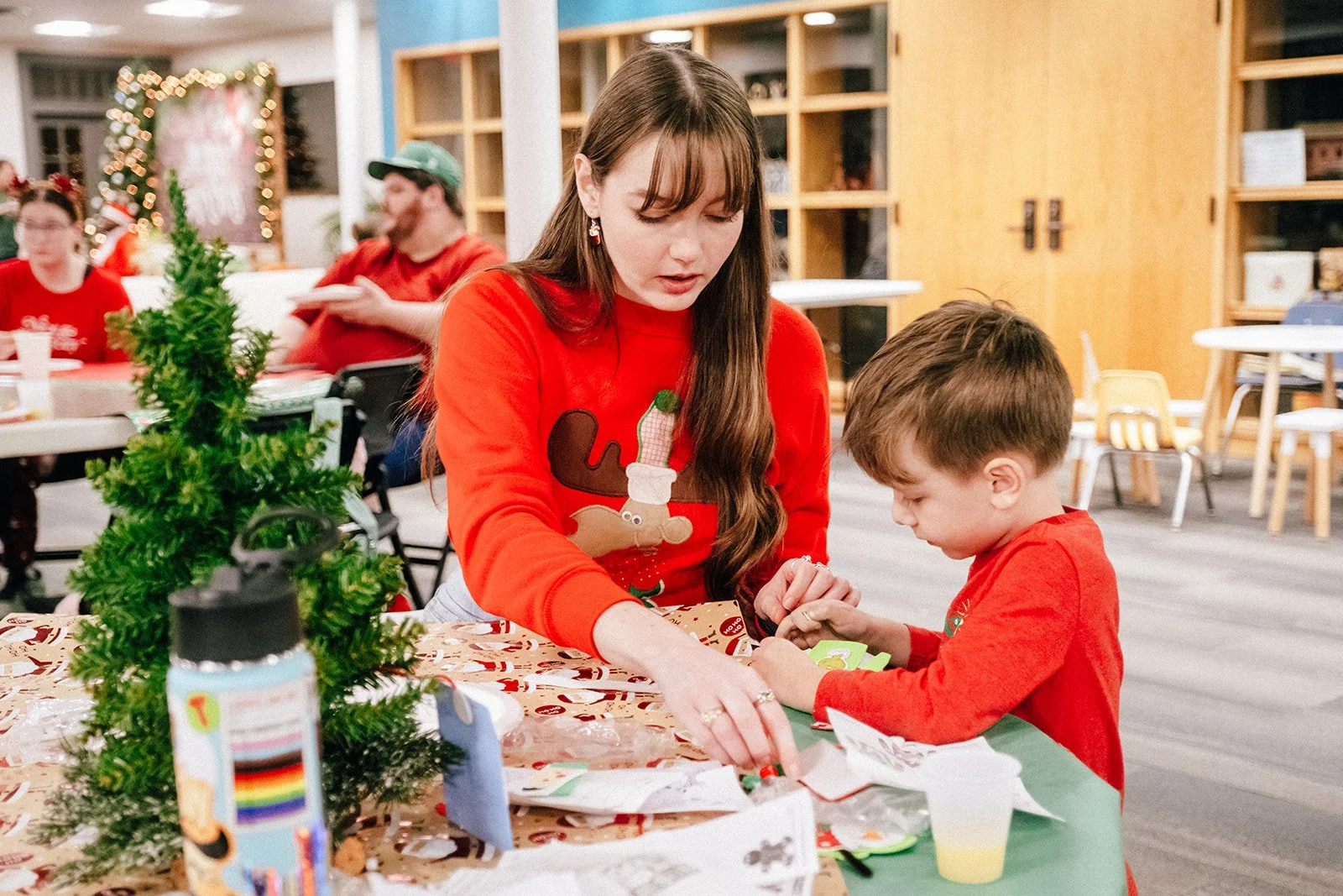 A young woman with brown hair sits at a table with a child helping him create a Christmas craft with paper and stickers.