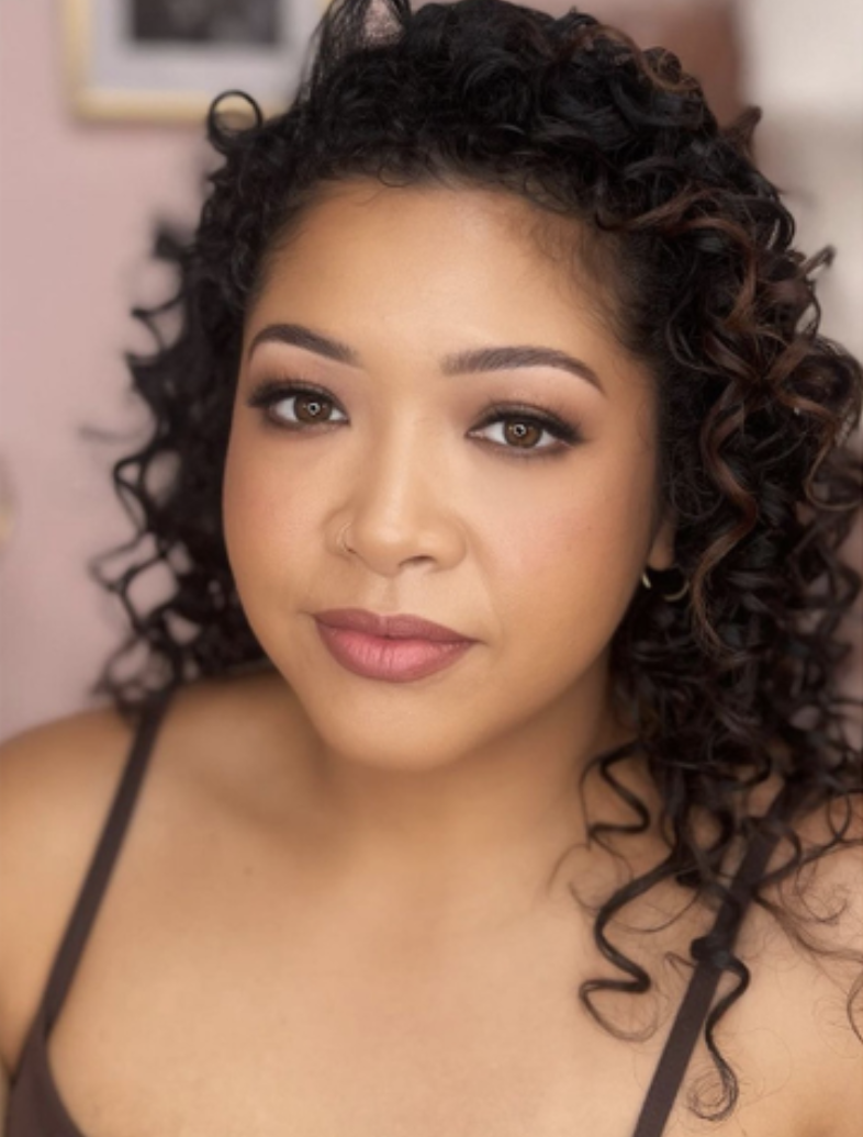 Close-up portrait of a young woman with curly dark hair, wearing makeup with defined eyebrows and natural lip color, and a nose ring, against a blurred background.