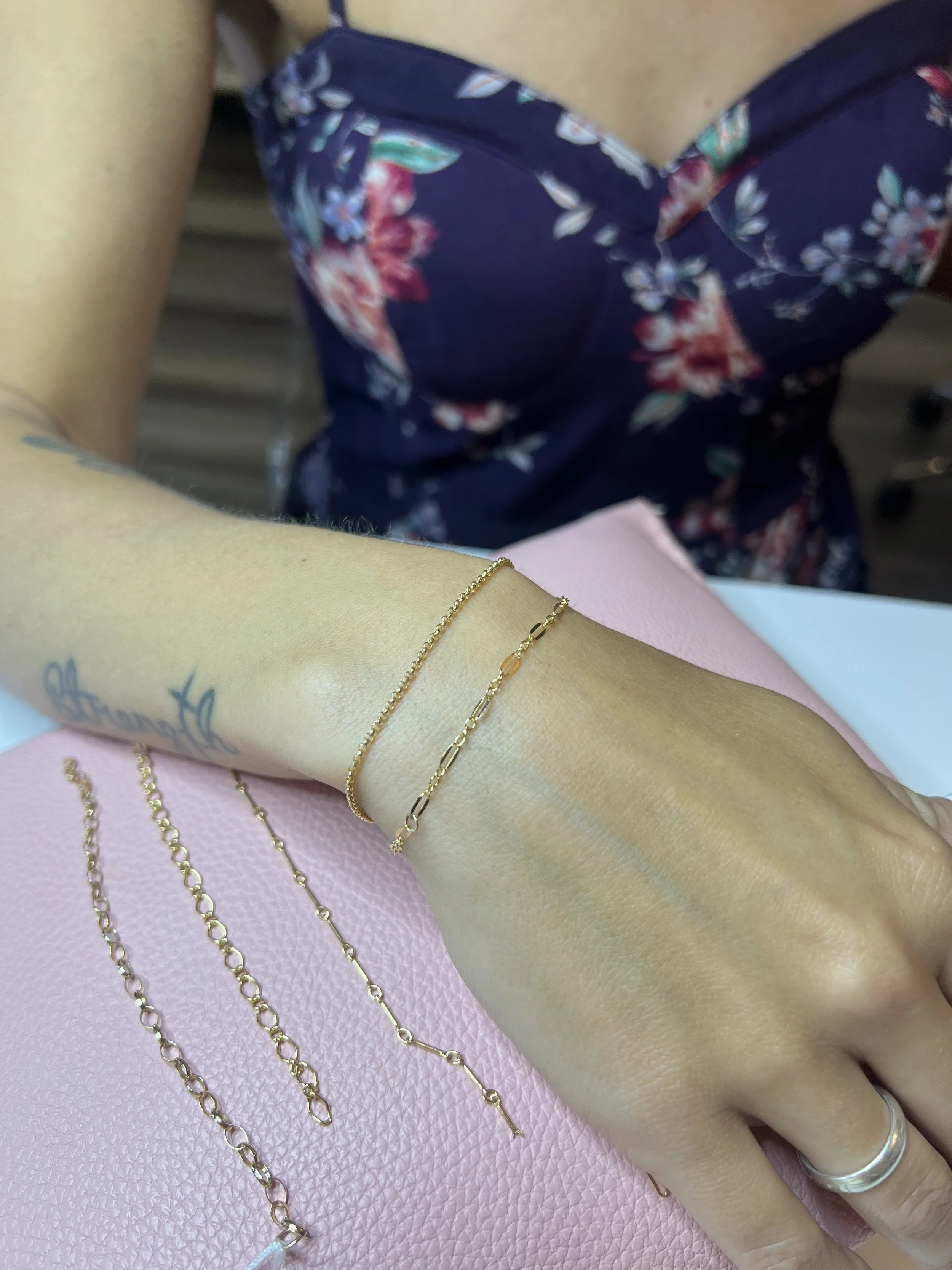 Person wearing two delicate gold bracelets on the wrist, with a floral dress and additional gold chains on a pink surface.