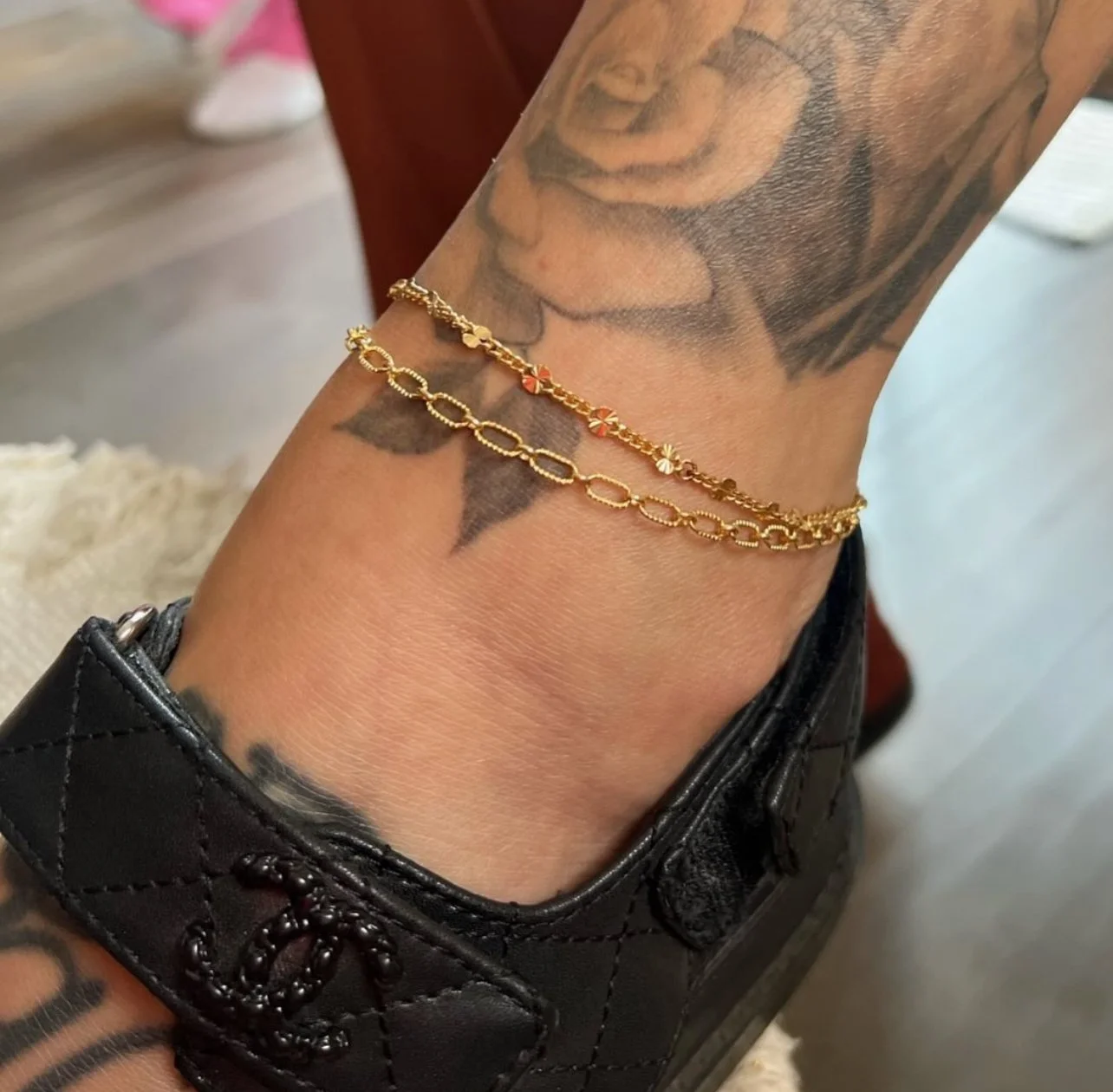 Close-up of a foot with rose tattoo, wearing two gold chain anklets, and black slip-on shoe with quilted pattern.