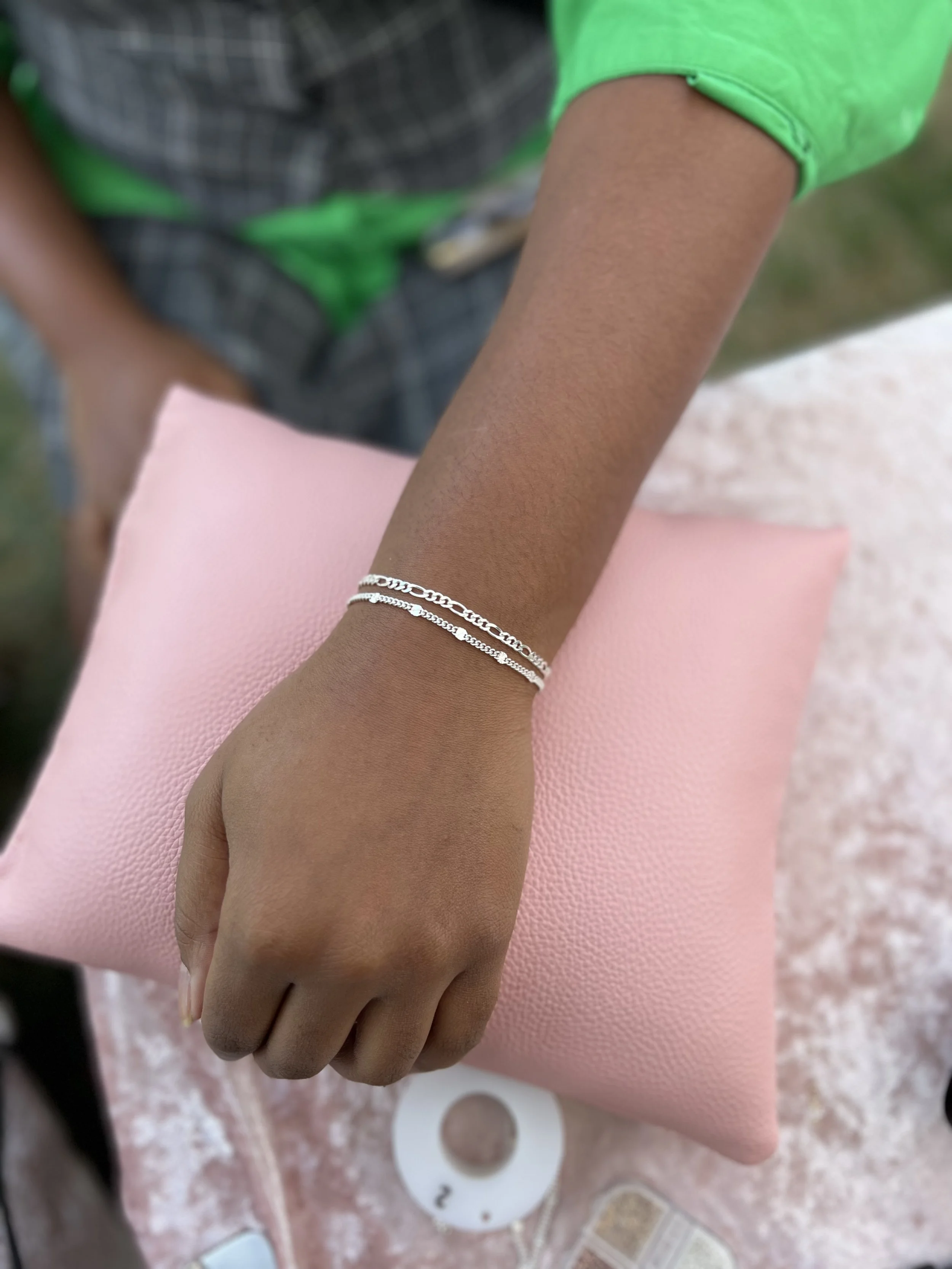 A person wearing a silver bracelet on their wrist, resting on a pink textured cushion. The person is wearing a green garment, and a few decorative or accessory items are visible in the background.