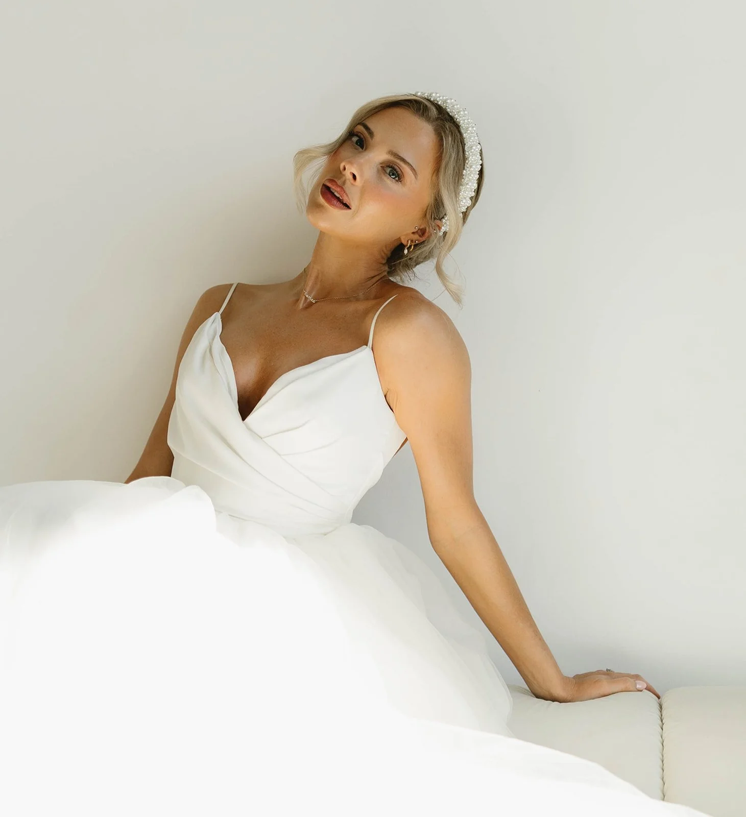 A woman in a white wedding dress with a pearl headband, sitting on a white couch against a plain wall, looking at the camera.