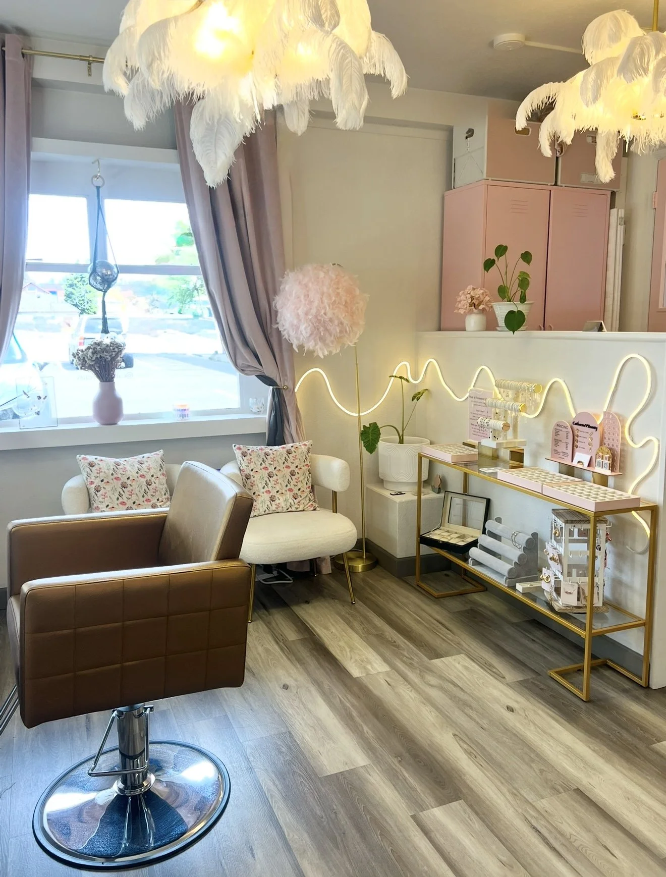A stylish hair salon interior with a brown salon chair, white armchair with floral pillows, pink cabinets, decorative plants, and feather-like light fixtures. A shelf with hair products is adorned with decorative lighting.