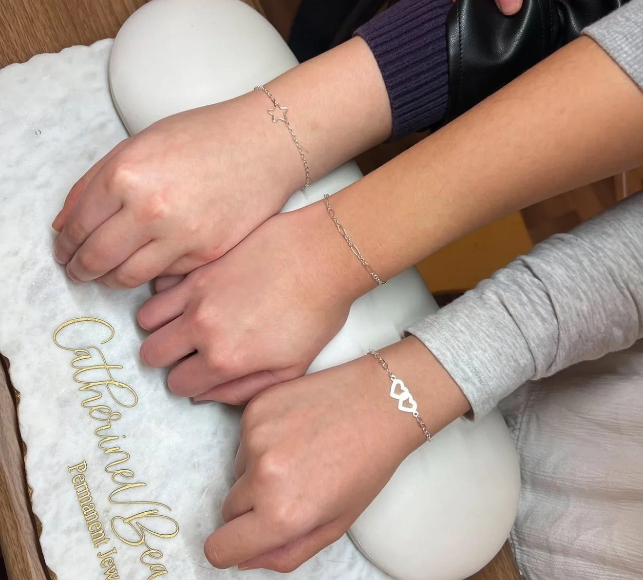 Three hands with silver bracelets on a marble surface labeled "Catherine Beauford Permanent Jewelry."
