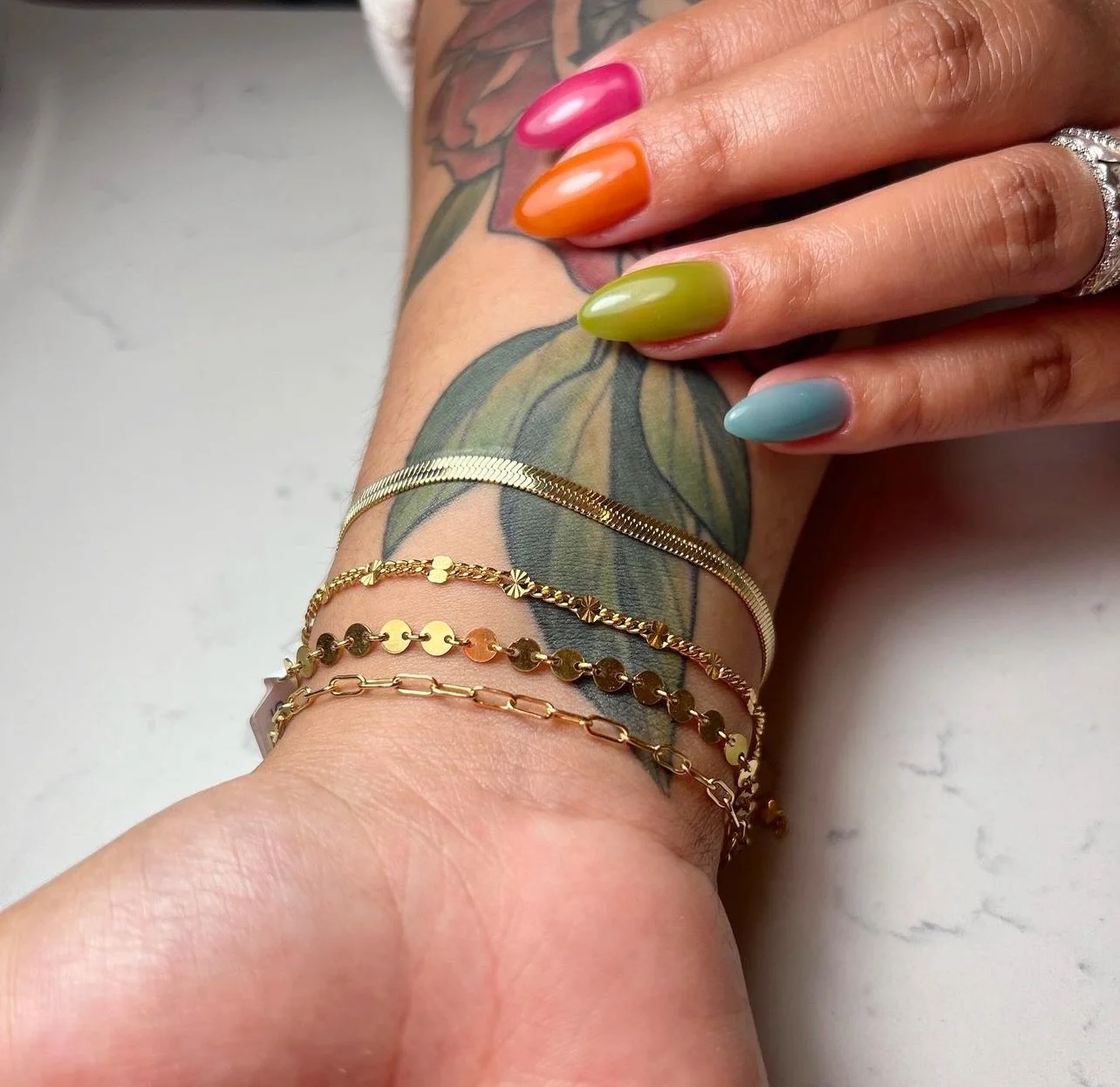 Close-up of a hand with multicolored manicured nails resting on a wrist adorned with gold bracelets, showcasing a floral tattoo on the forearm.