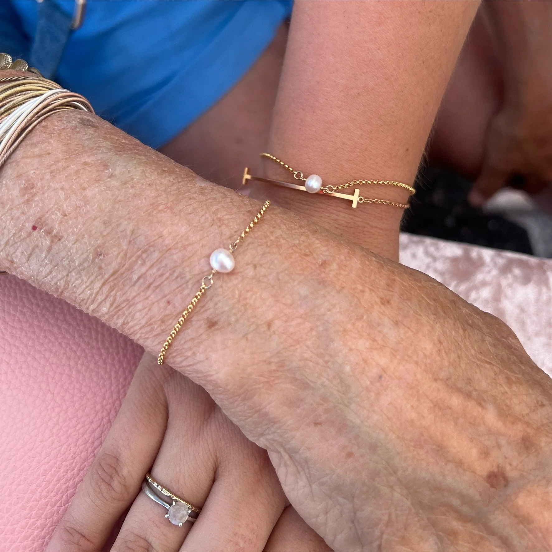 Two wrists with gold chain bracelets featuring pearls, and one hand wearing a ring with a clear stone set on a band with smaller stones.