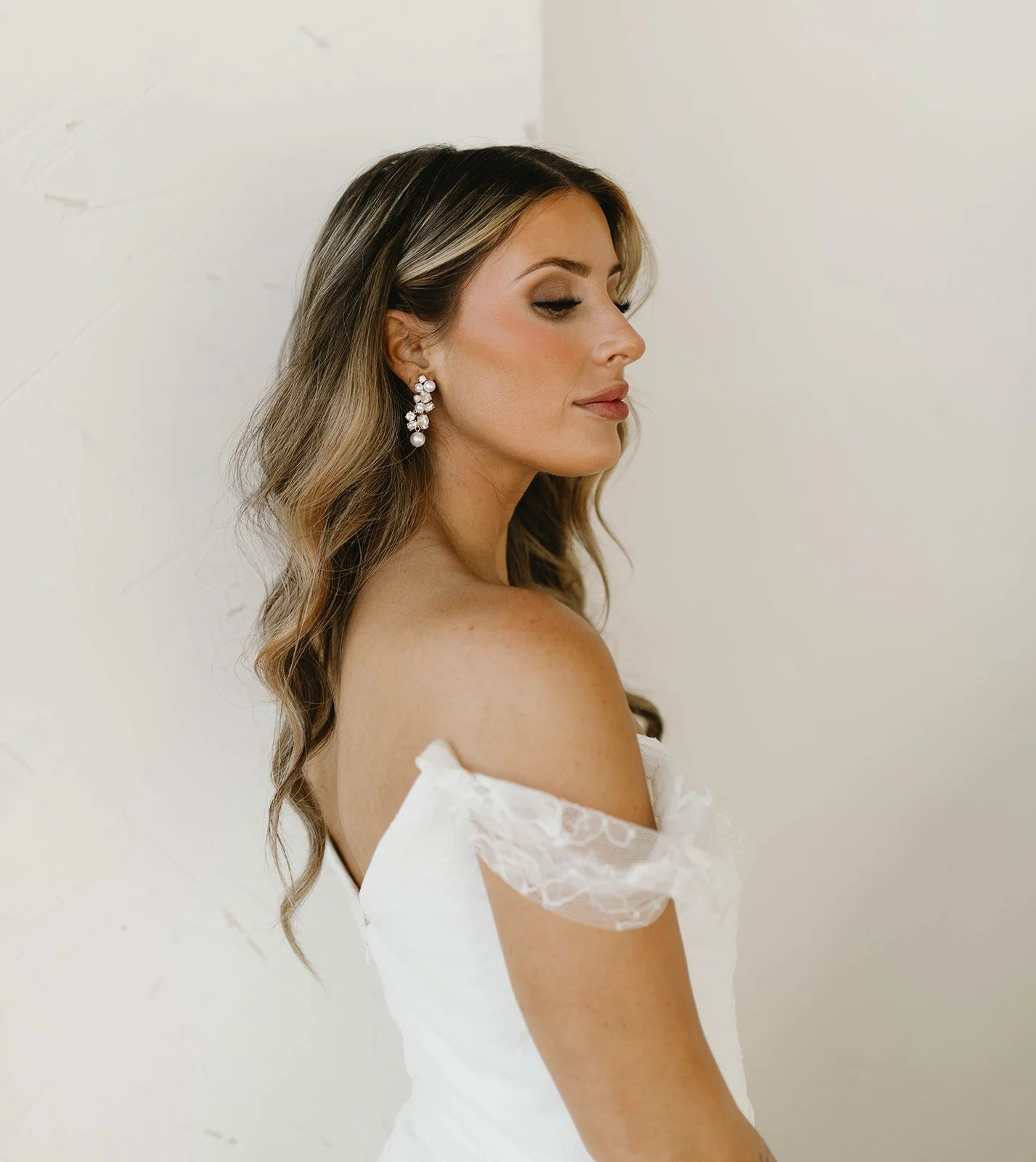 Close-up of a woman with long wavy hair, wearing pearl earrings and a white off-shoulder dress, standing against a plain white background.