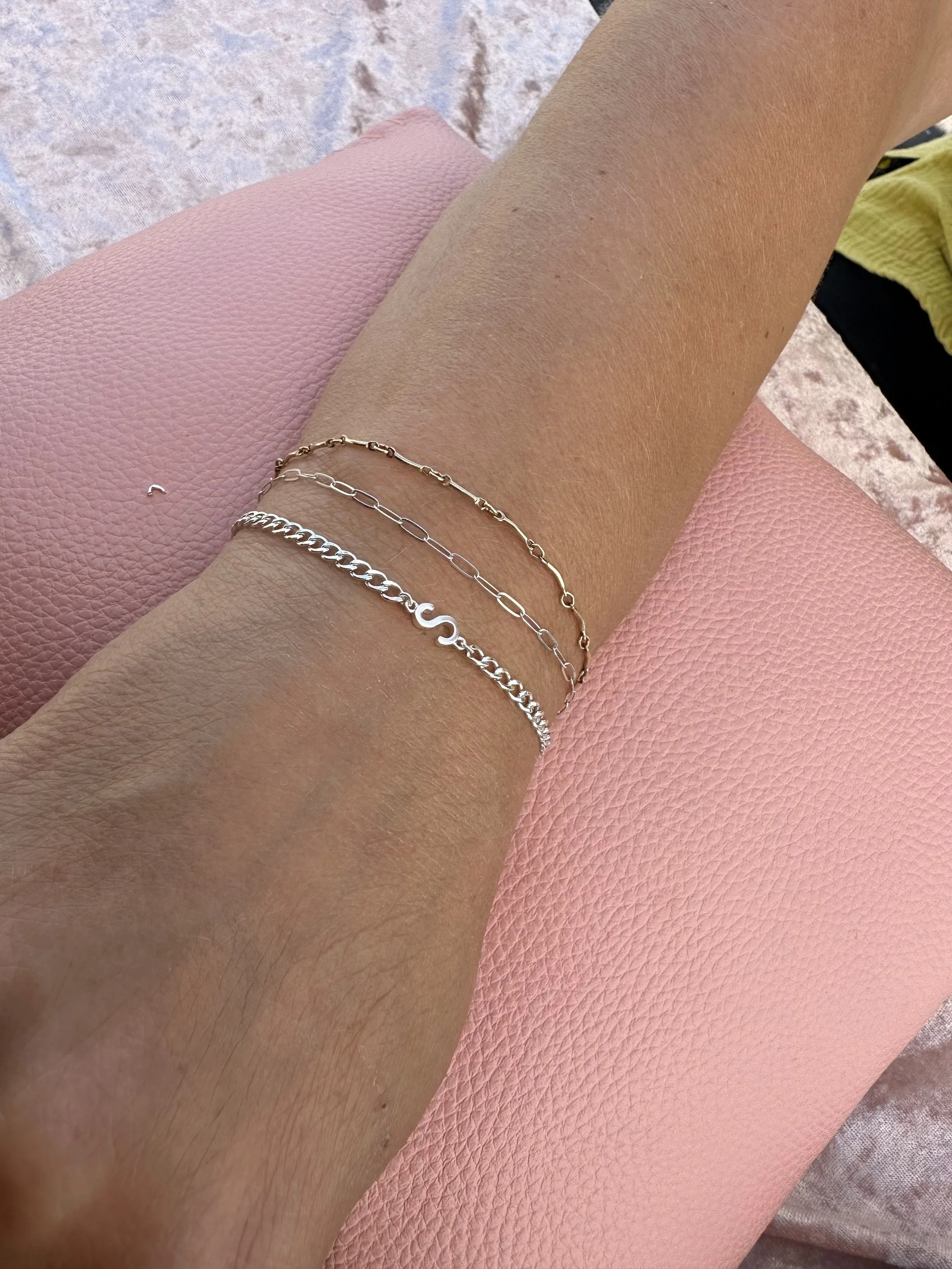 A close-up view of an arm wearing three delicate chain bracelets, with a combination of silver and gold tones, resting on a textured pink cushion.