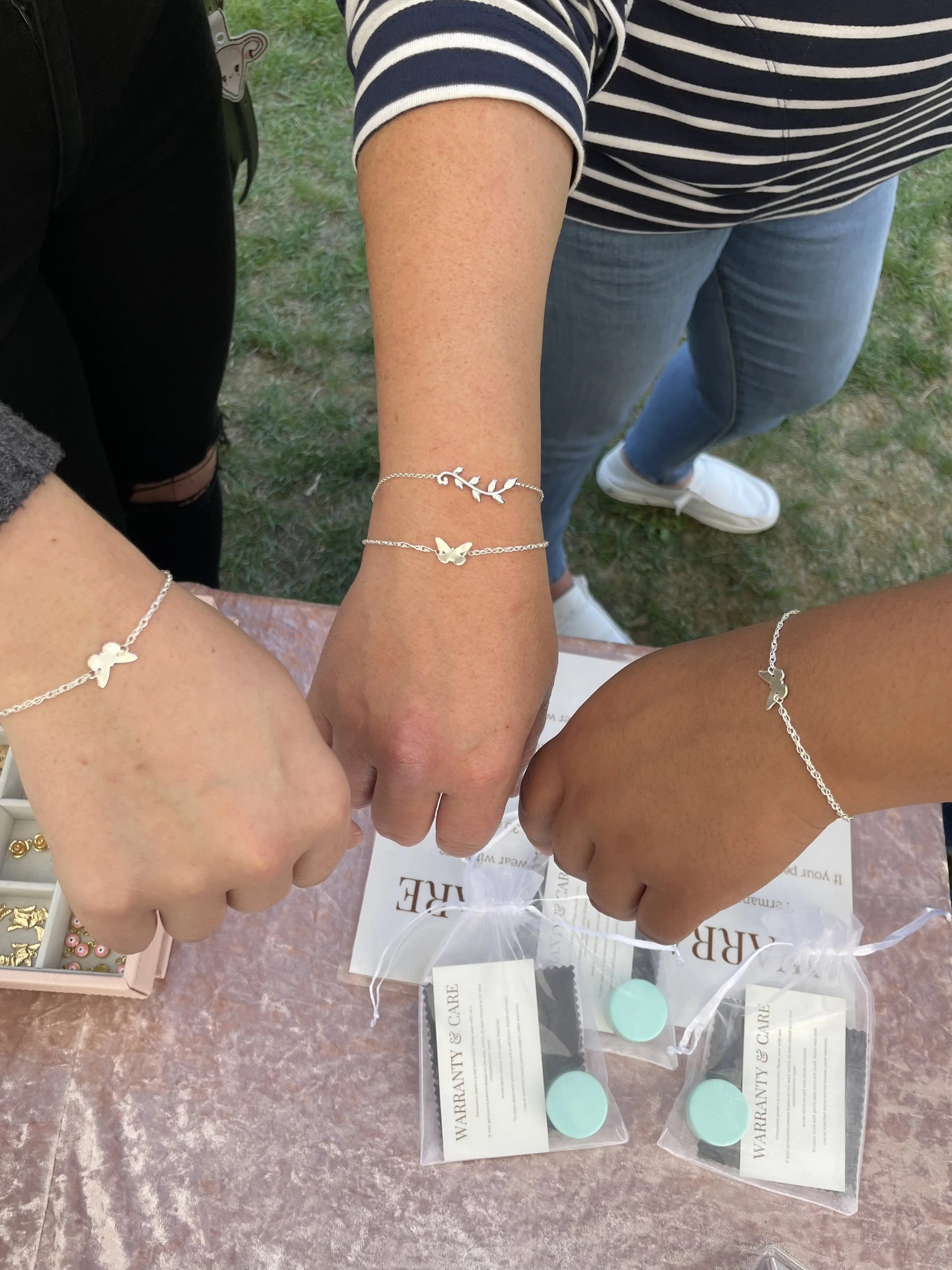 Three people displaying their arms wearing delicate silver bracelets with charm designs, including a butterfly and foliage. They're standing over a table with warranty cards and jewelry care pouches.