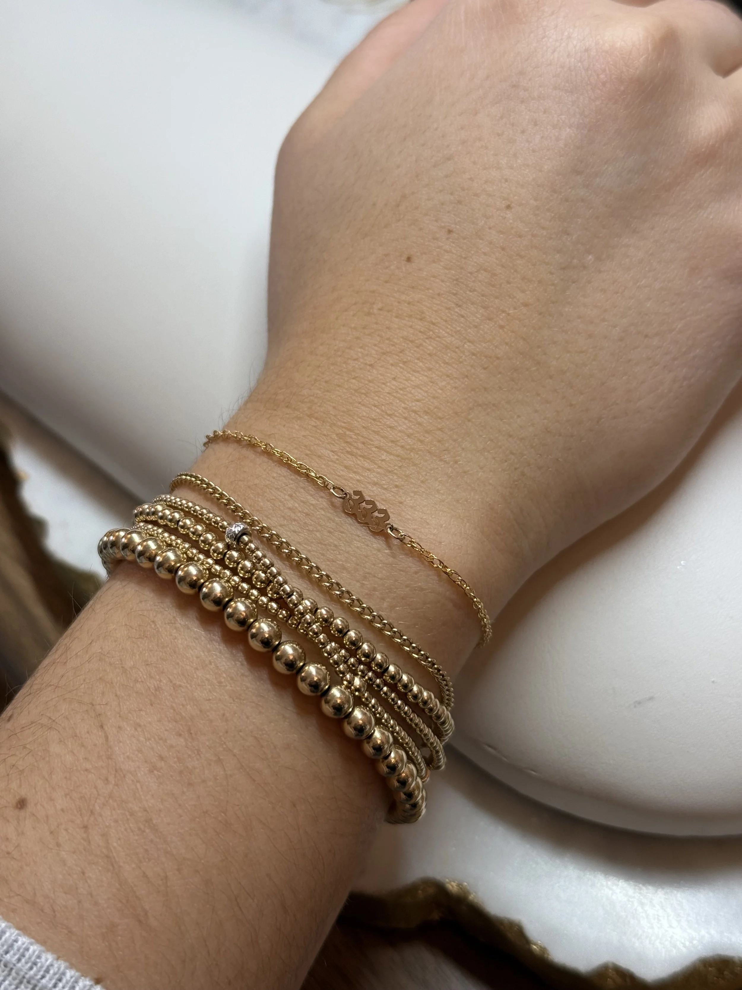 Wrist adorned with multiple gold bracelets, including beaded and chain designs, on a person's arm resting on a white surface.