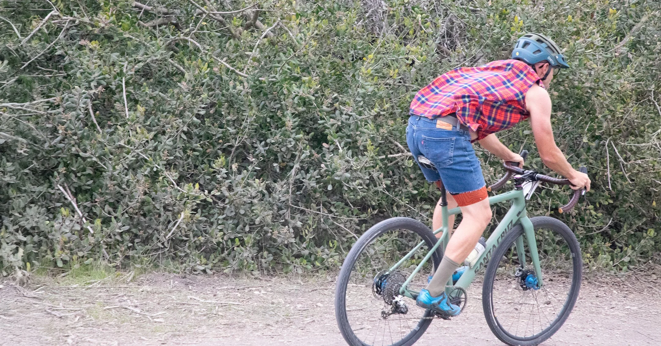 A man riding a mountain bike on a dirt trail surrounded by bushes, wearing a blue helmet, sleeveless plaid shirt, denim shorts, and sneakers.