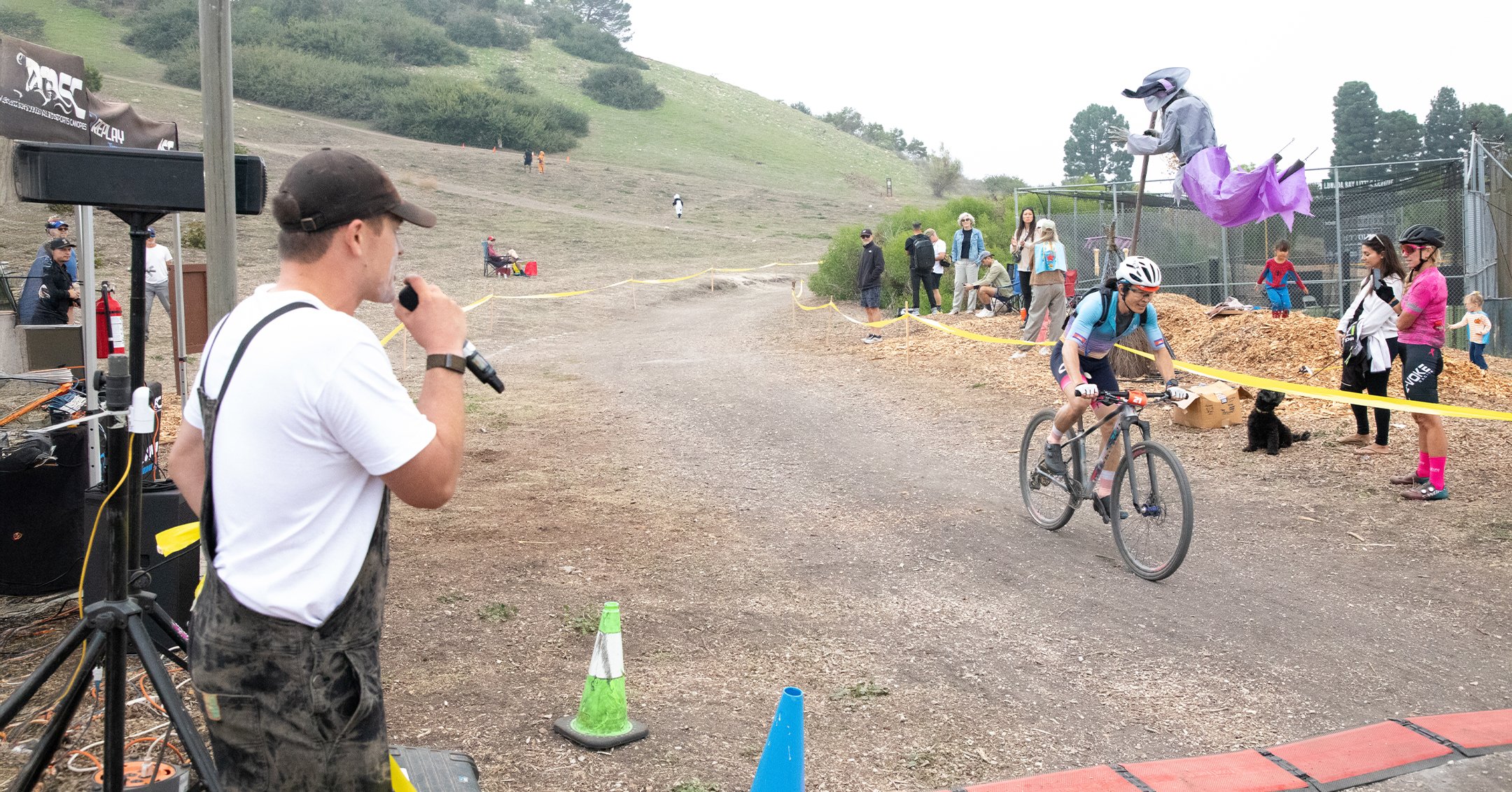 A cyclist crossing the finish line at a race, while a man with a microphone announces, and spectators watch along yellow tape on a dirt path in a hilly outdoor setting.