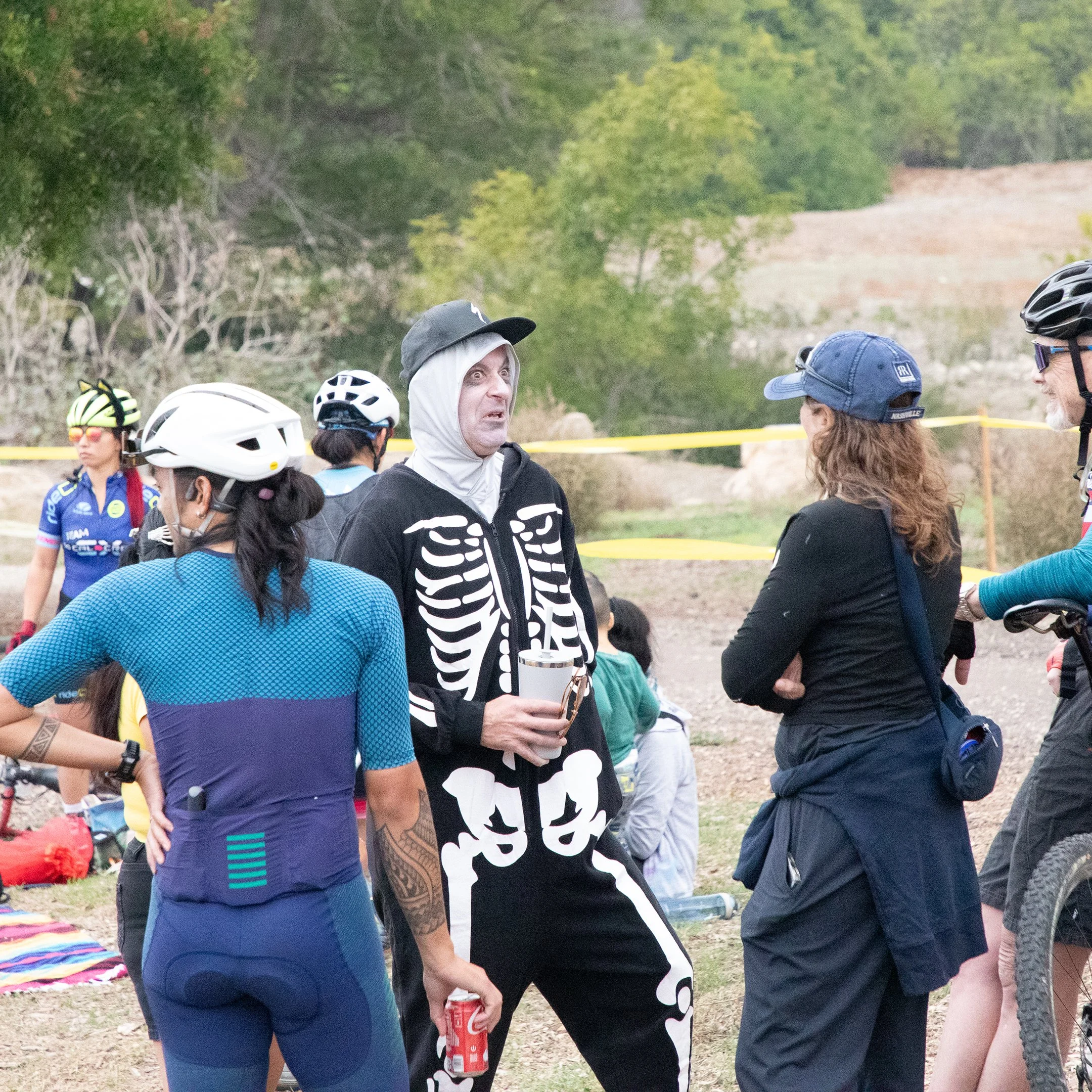 Group of people, some wearing cycling gear and helmets, gathered outdoors in a park-like setting with trees and rocky terrain in the background.