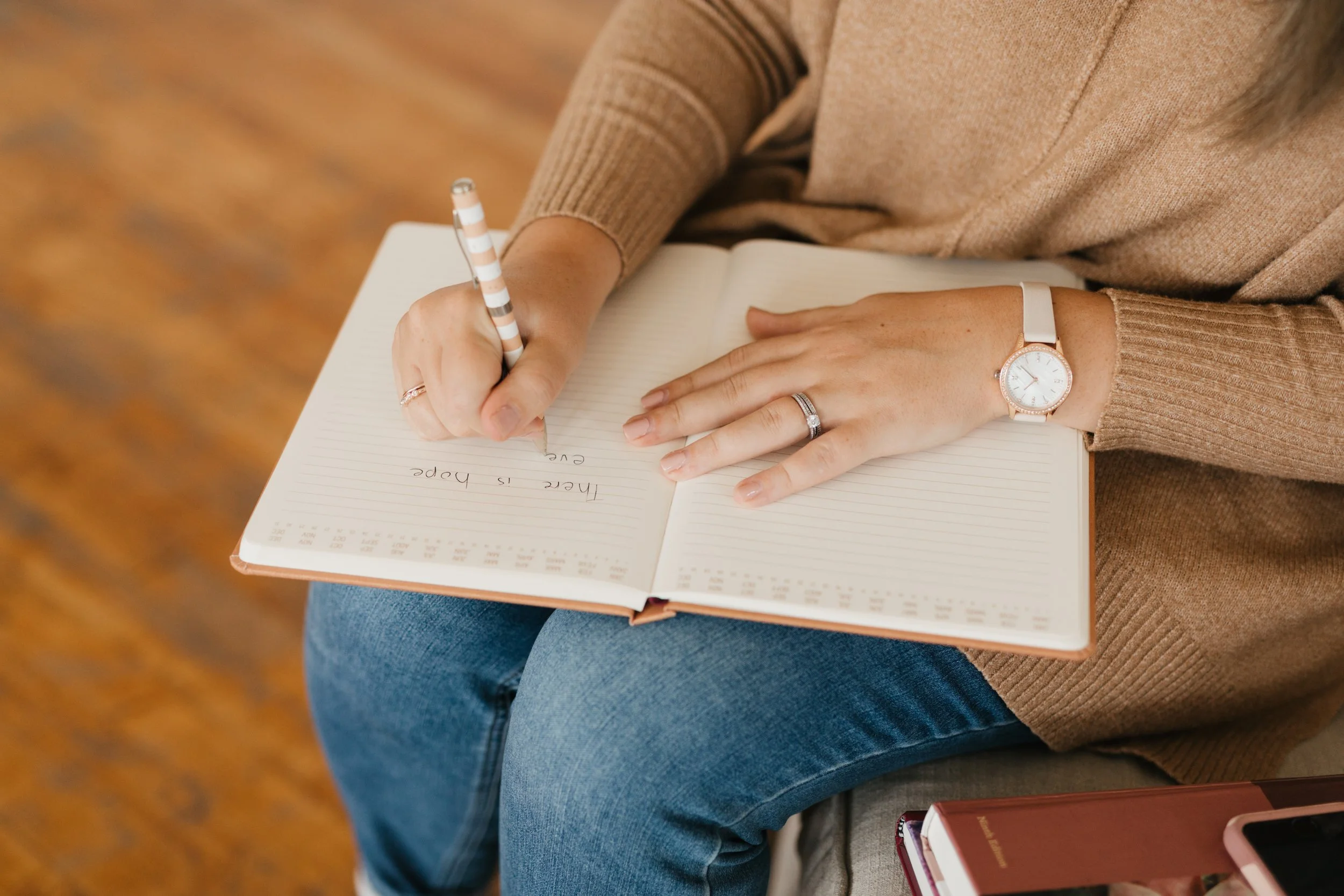A person wearing a beige sweater, jeans, and a watch is sitting and writing in a lined notebook with a striped pen. The person has rings on their fingers and is seated on a chair with a brown carpeted floor visible in the background.