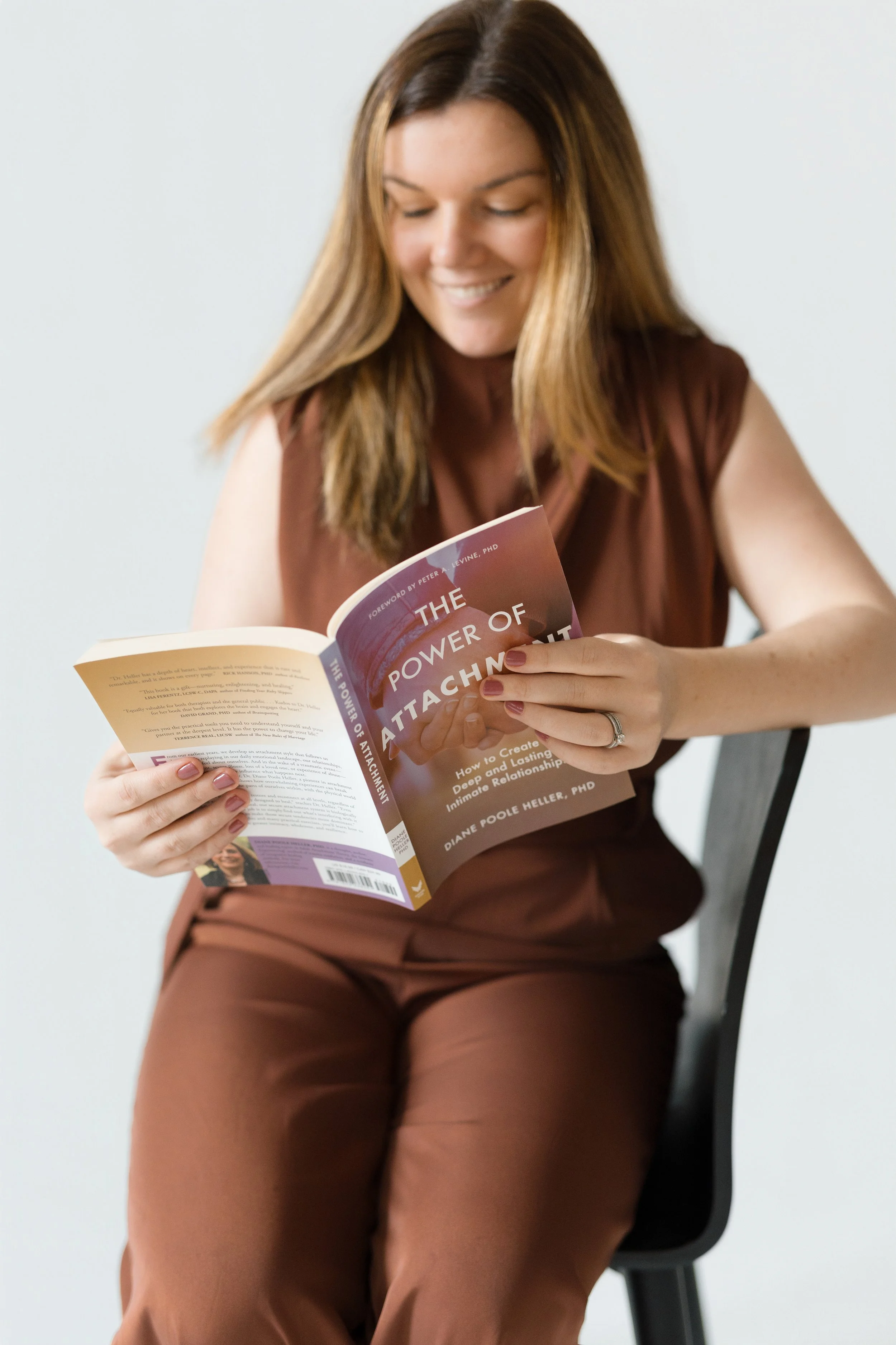 A woman with long, wavy brown hair and light skin is sitting on a black chair, smiling while reading a book titled 'The Power of Attachment' by Diane Poole Heller, PhD, against a plain white background.