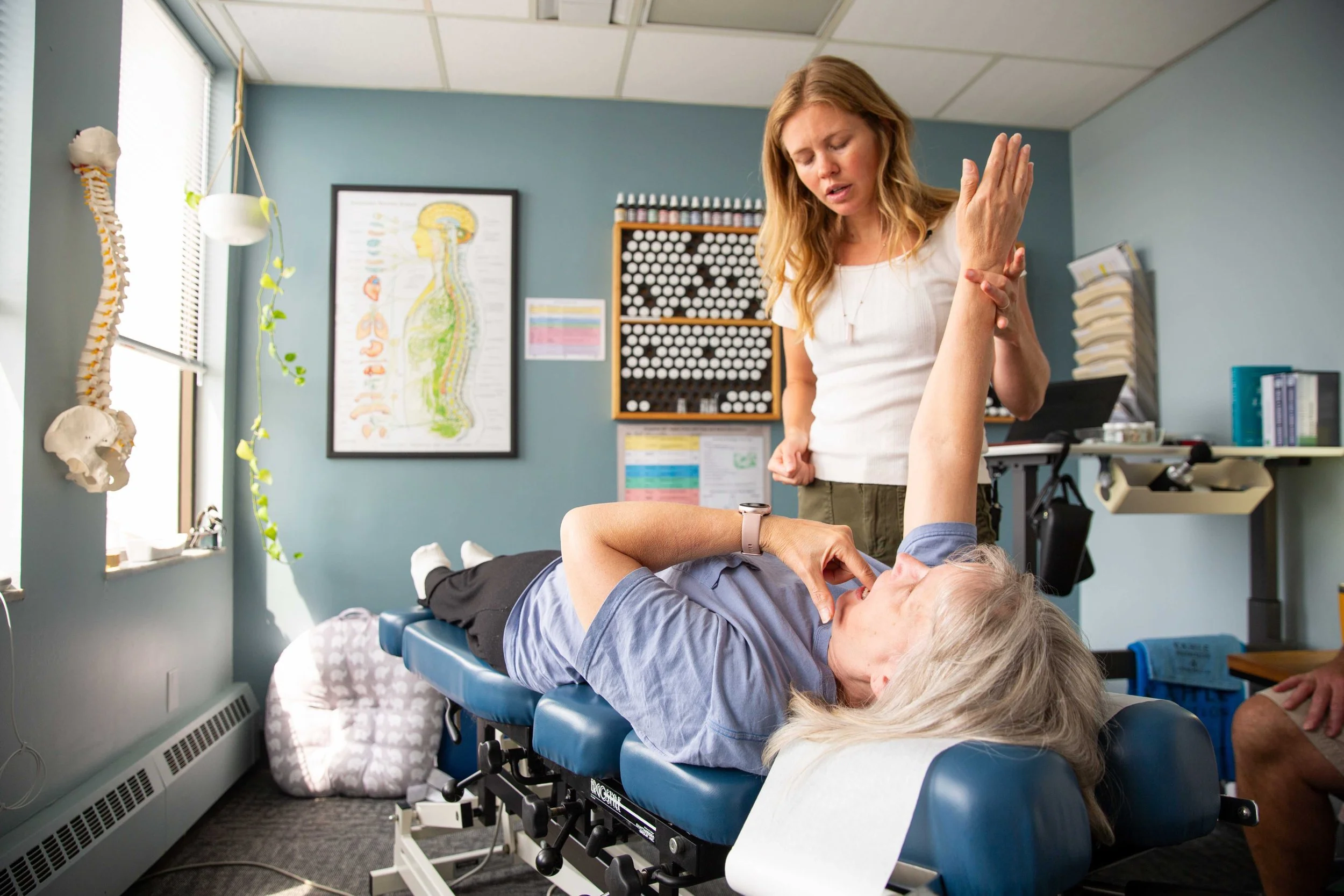 A chiropractor adjusts a patient's arm while the patient is lying on a chiropractic table in a medical office with anatomical charts and files in the background.