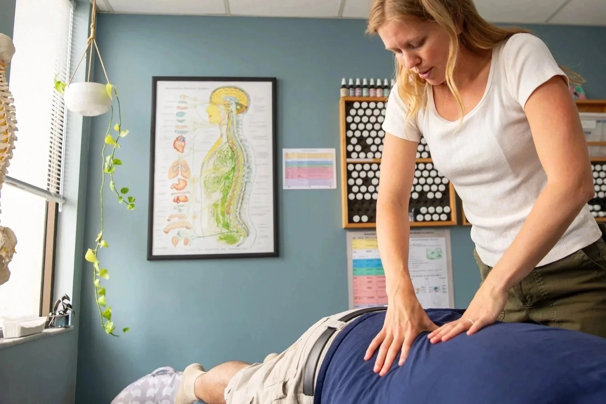 Chiropractor adjusting back of patient lying face down on treatment table in clinic, with anatomical spine chart and drug storage in background.