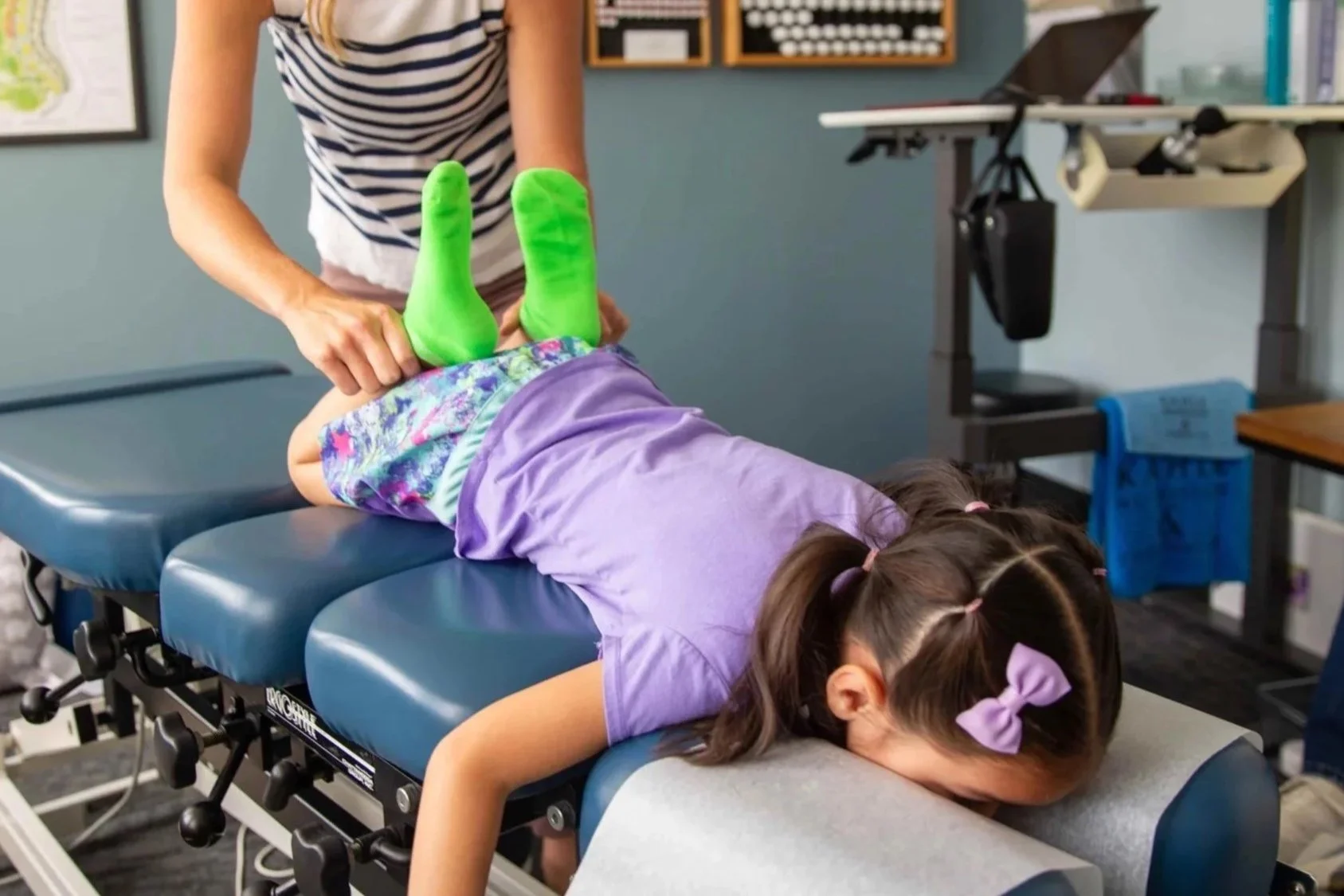 Child lying face down on a therapy table with legs raised and supported by a therapist, wearing colorful clothing and green socks, in a medical or therapy room.