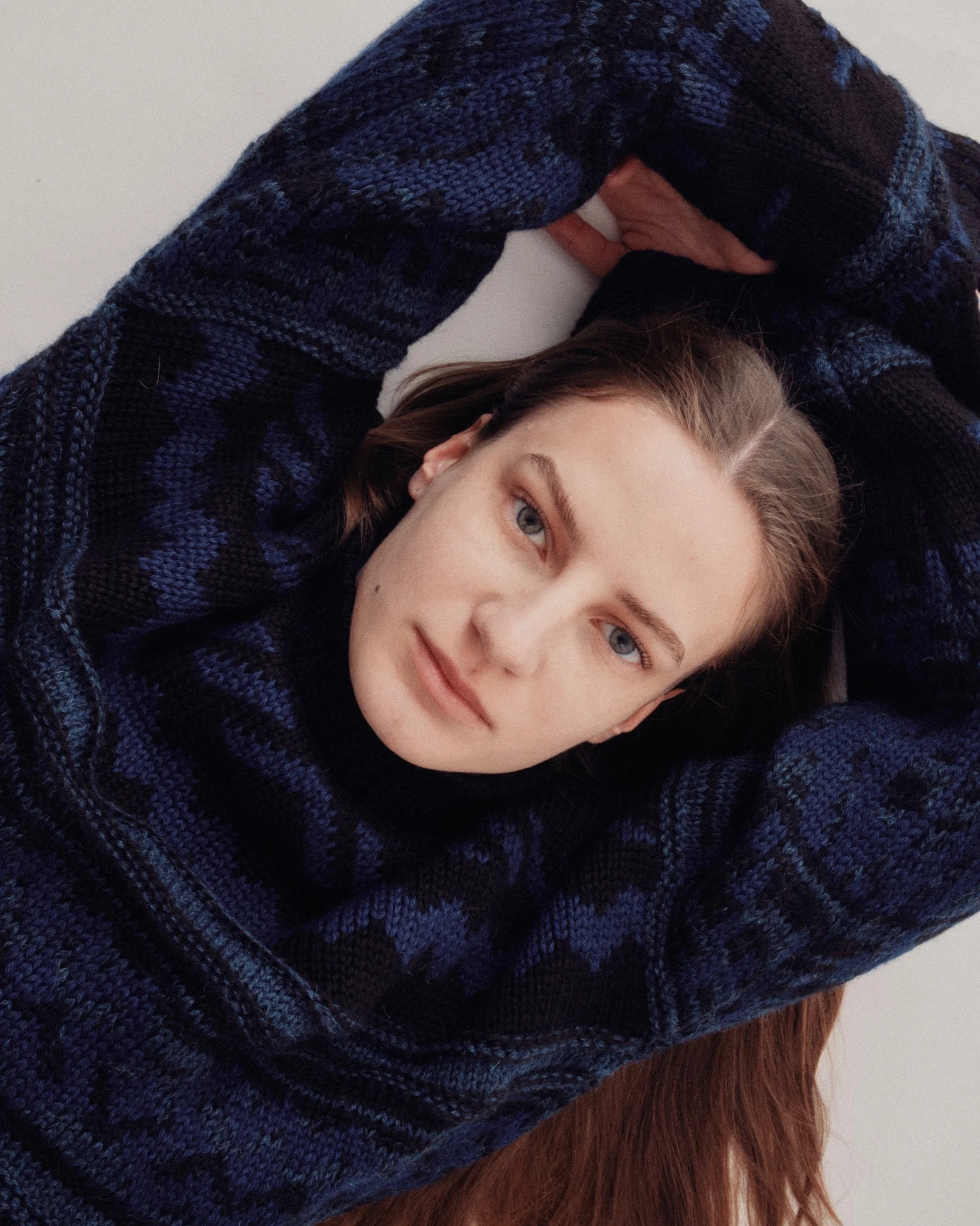 A woman with long brown hair and blue eyes lying down with arms behind her head, wearing a blue and black patterned sweater.