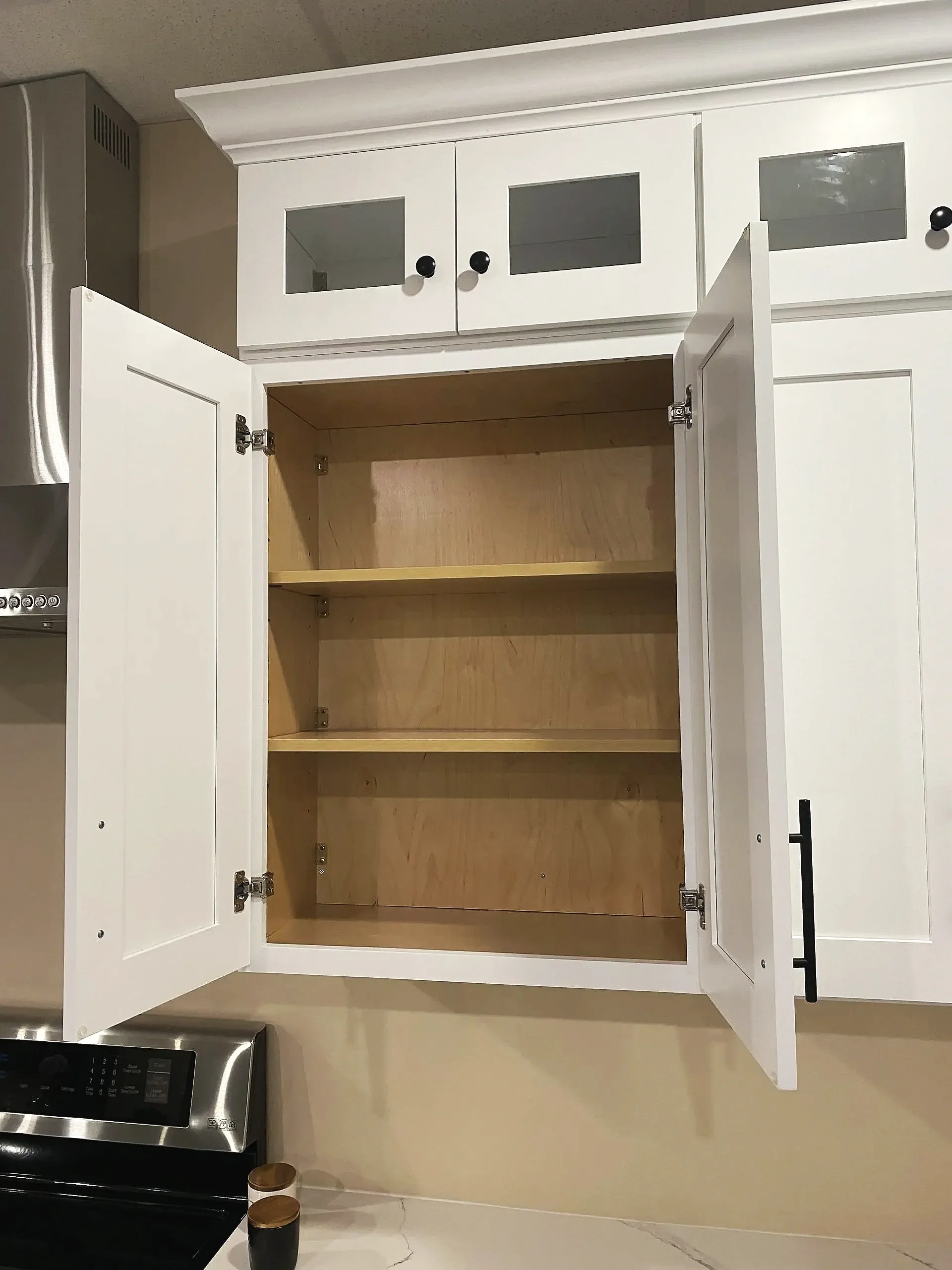 Empty white kitchen cabinet with open doors showing three wooden shelves inside, located above a stove, with a cream-colored wall behind.