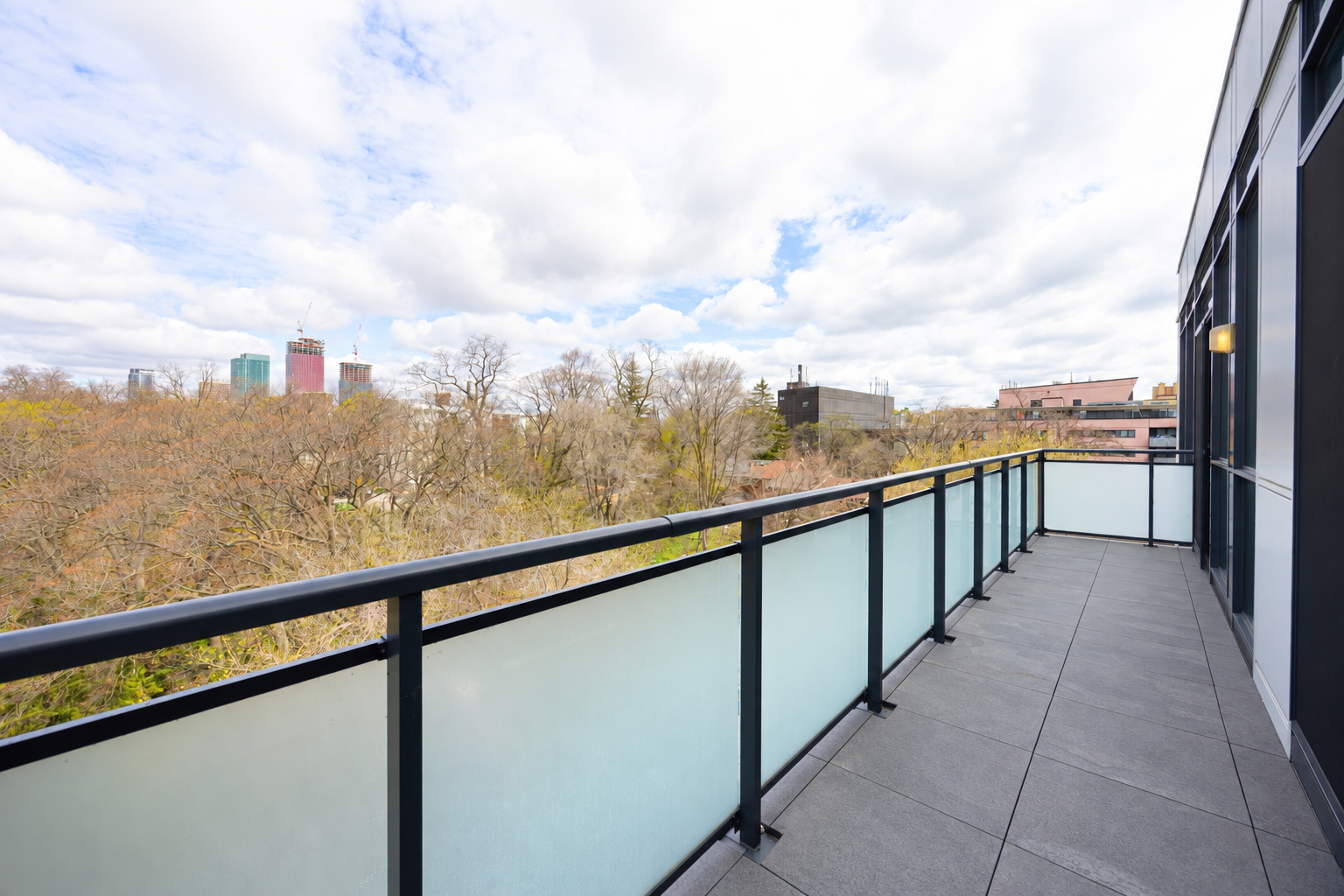 Empty condo balcony with gray tiled floor, black metal railing, frosted glass privacy screen, brick wall, and green trees in the background.