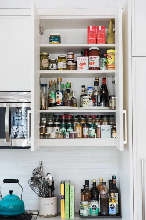 Open kitchen cabinet with shelves filled with jars, bottles, and boxes of food and condiments, next to a microwave oven and a blue kettle.