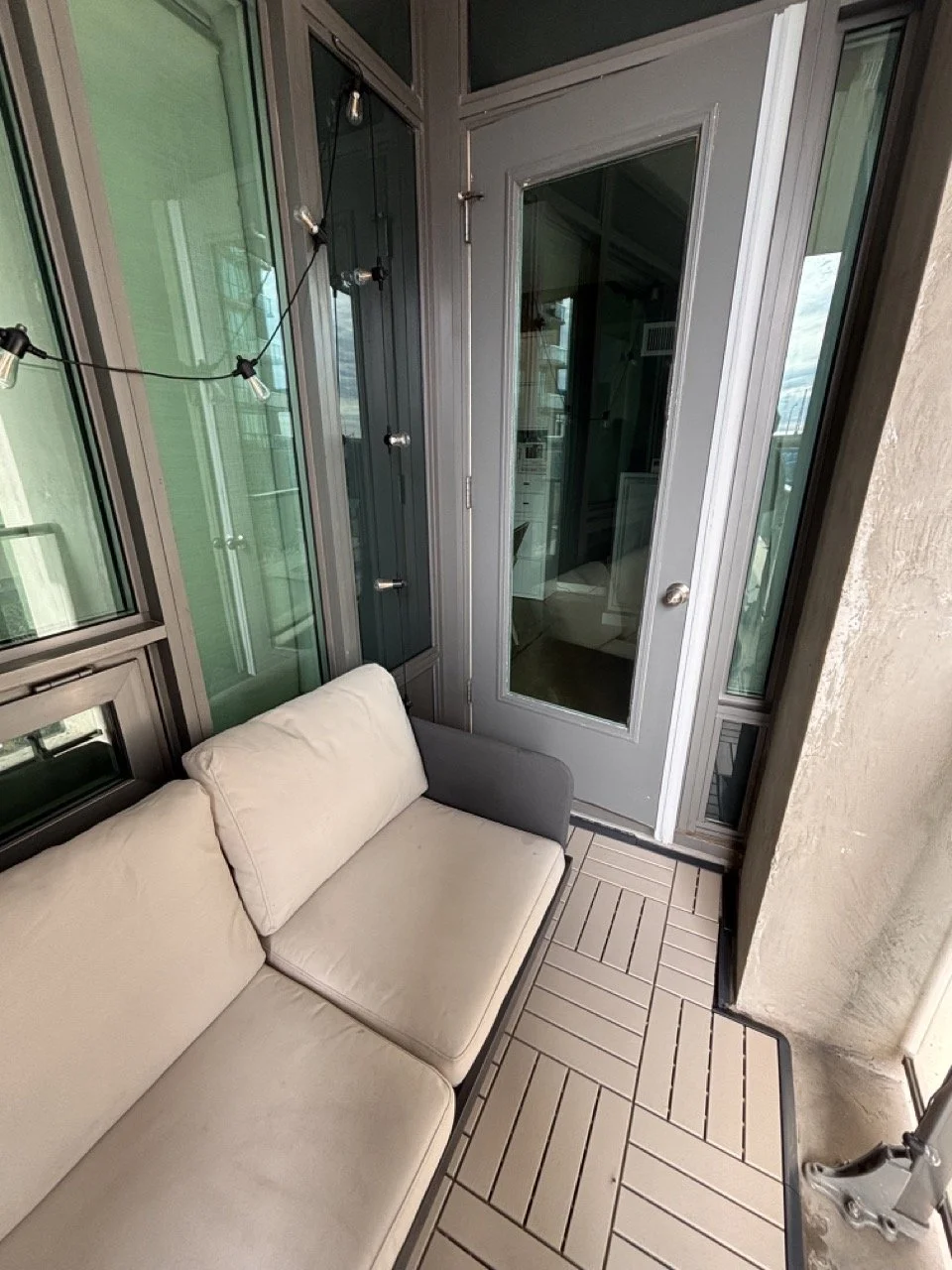 Small clean balcony in Liberty Village with a beige couch, glass door, string lights, and window panels.