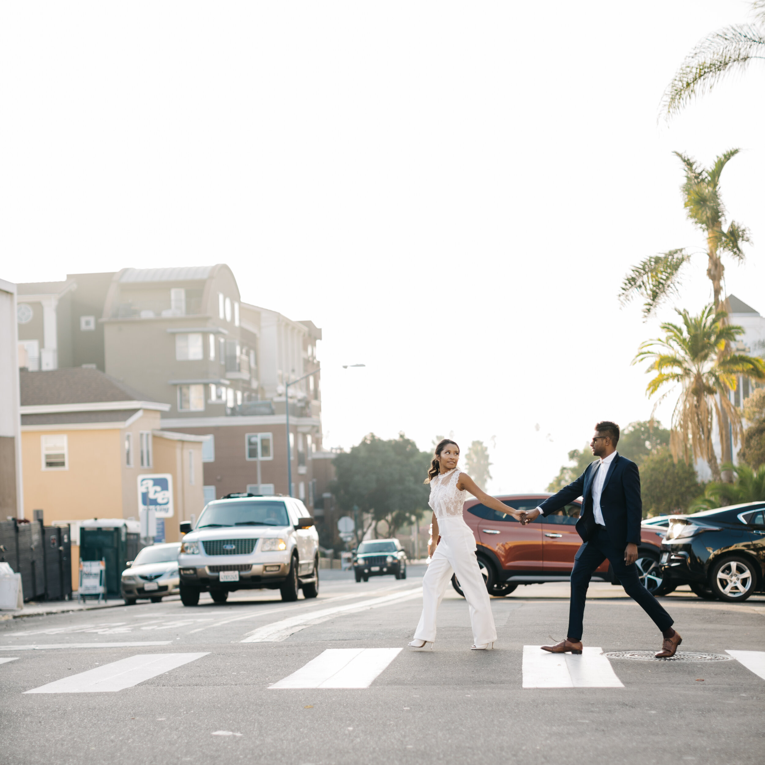 A couple holding hands and walking across a crosswalk in a suburban neighborhood with modern houses and palm trees, during daytime.