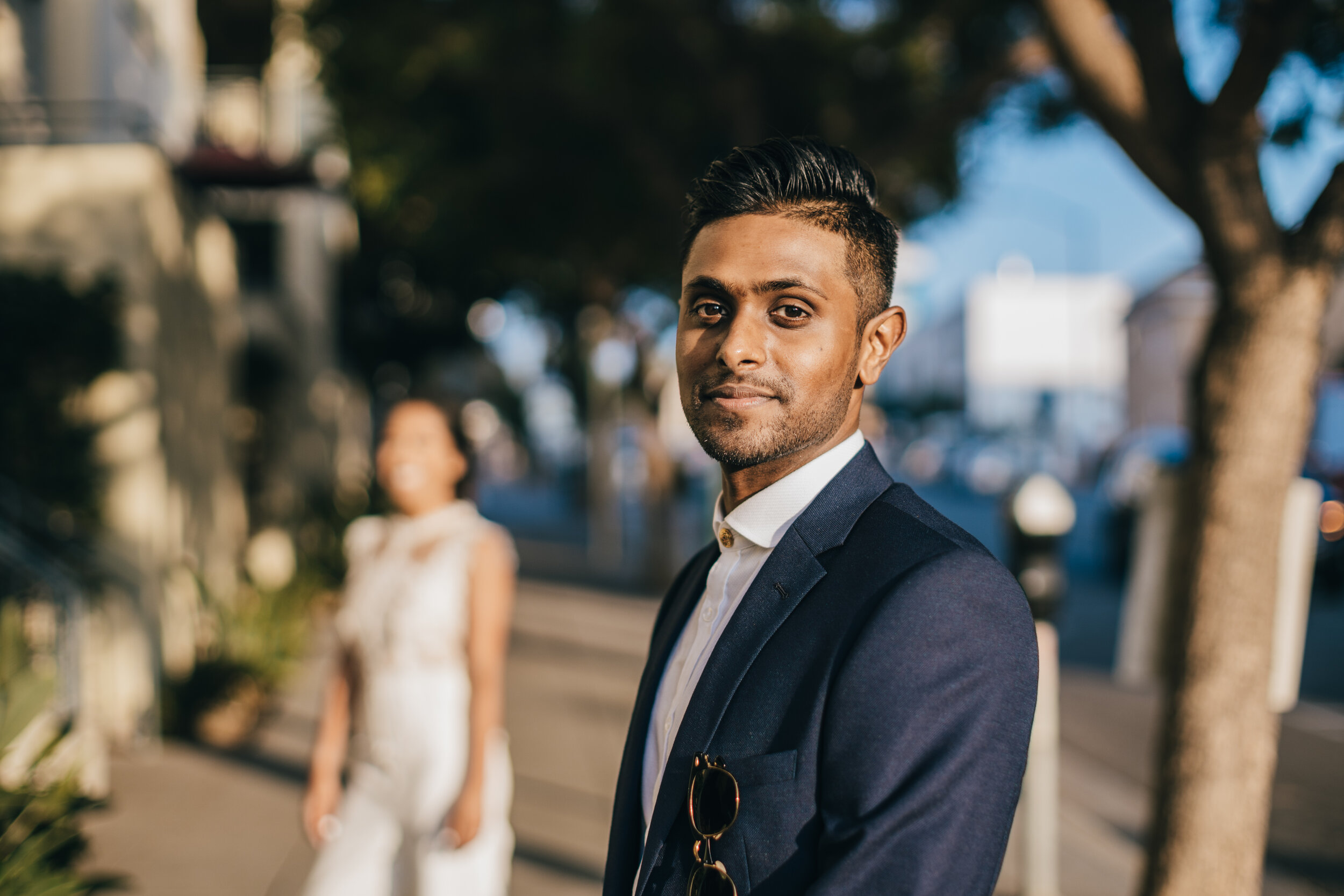 A man dressed in a navy blazer and white shirt, with sunglasses hanging from his blazer, standing outdoors with trees and a blurred background of people, smiling slightly and looking at the camera.