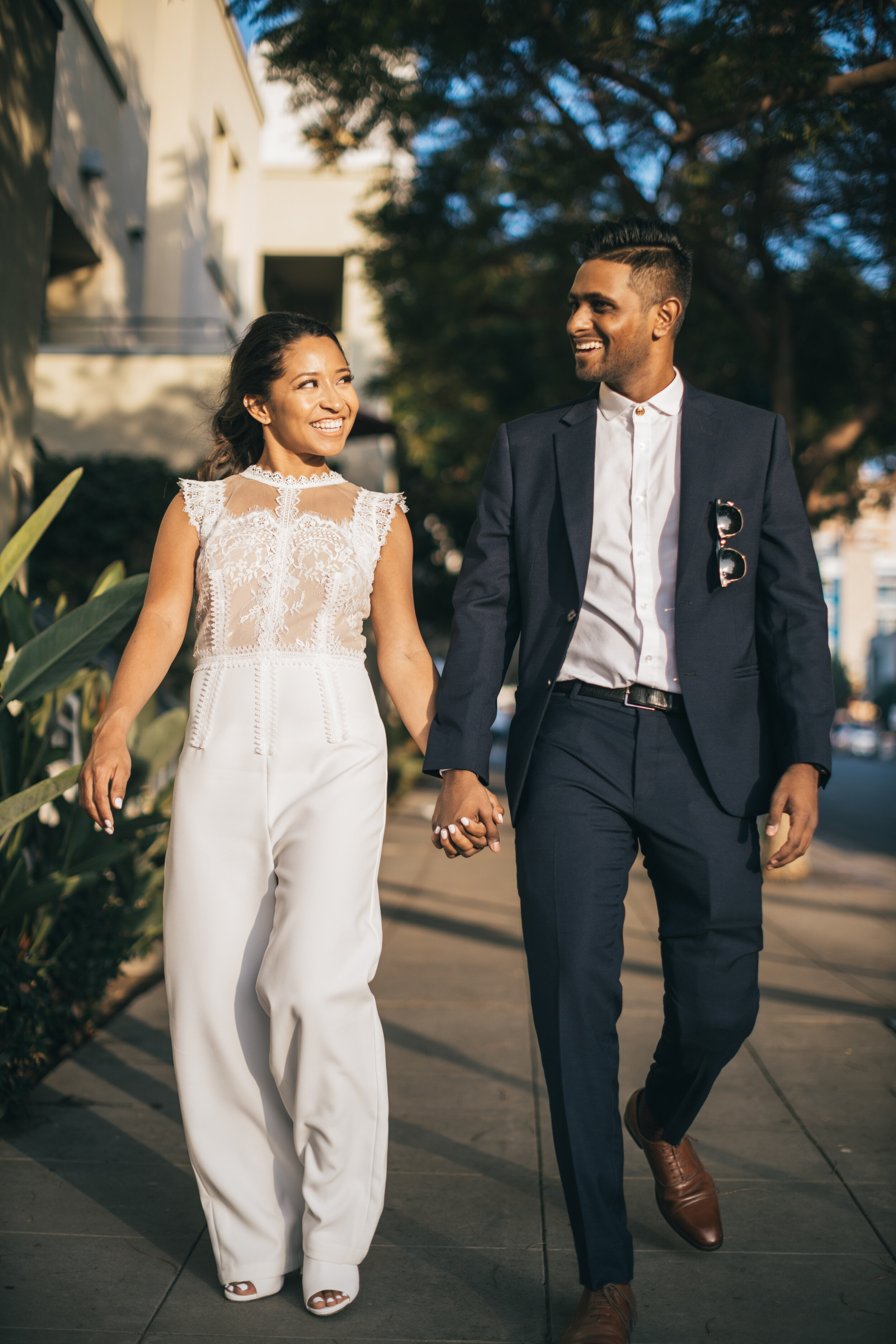 A smiling woman in a white lace top and white pants walking hand-in-hand with a man in a navy suit and white shirt outdoors in a sunny urban area.